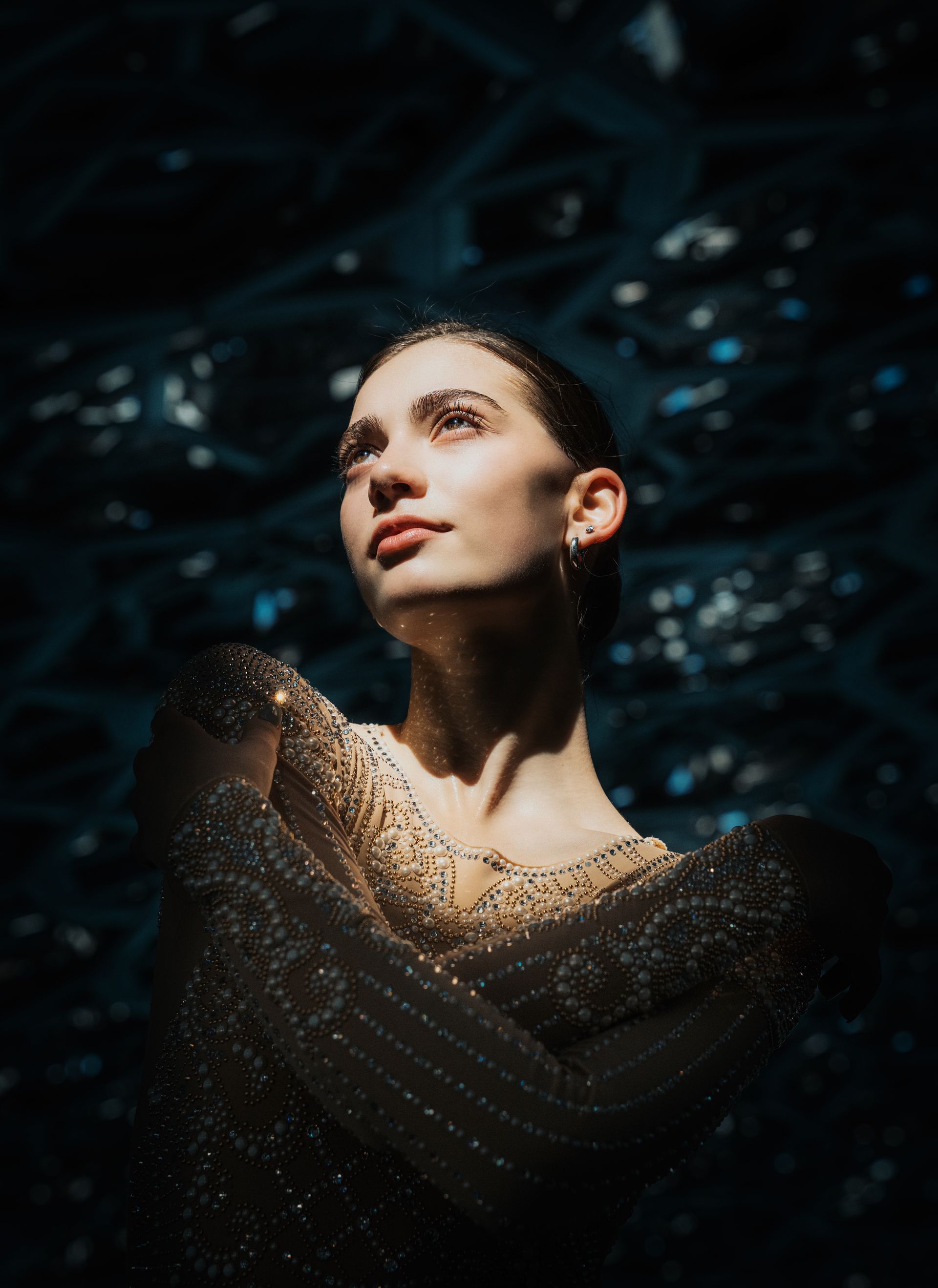 Woman in shimmering dress, arms crossed, looking up with a soft expression. Dark, abstract background.