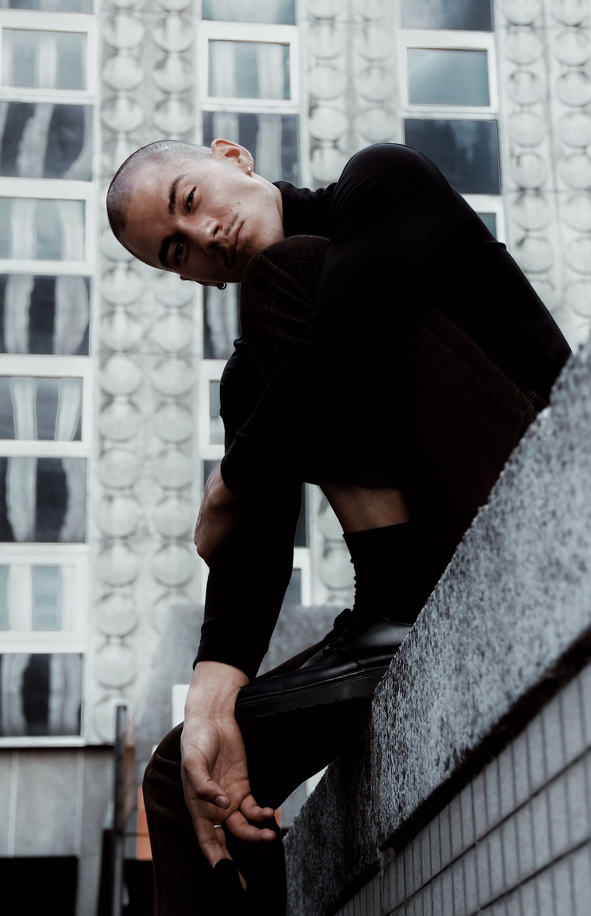 Man in black turtleneck, sitting on a concrete ledge, looking at the camera. Building in the background.