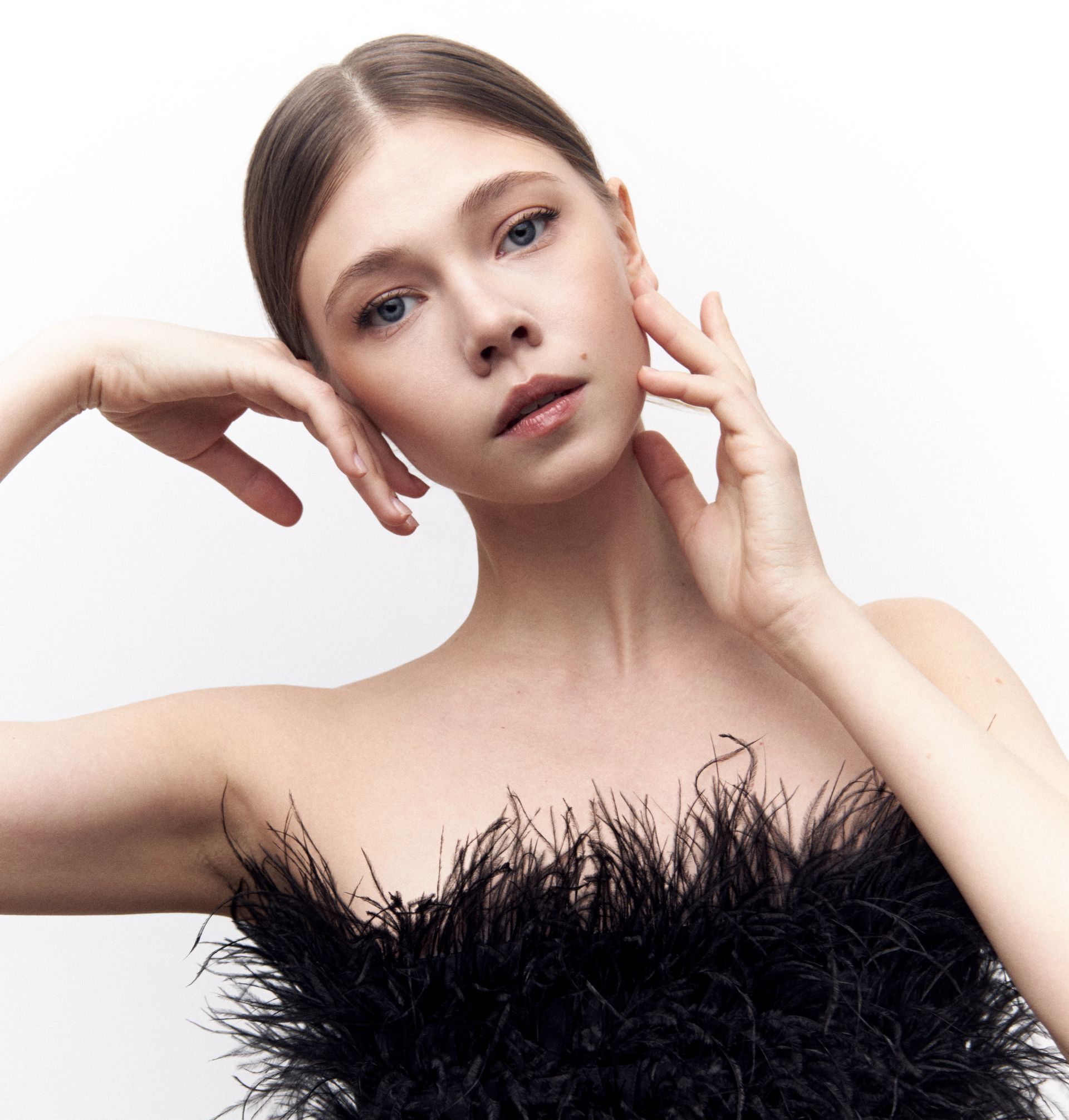 Woman in a black, feathered strapless top, hands near face, gazing upward with a neutral expression.