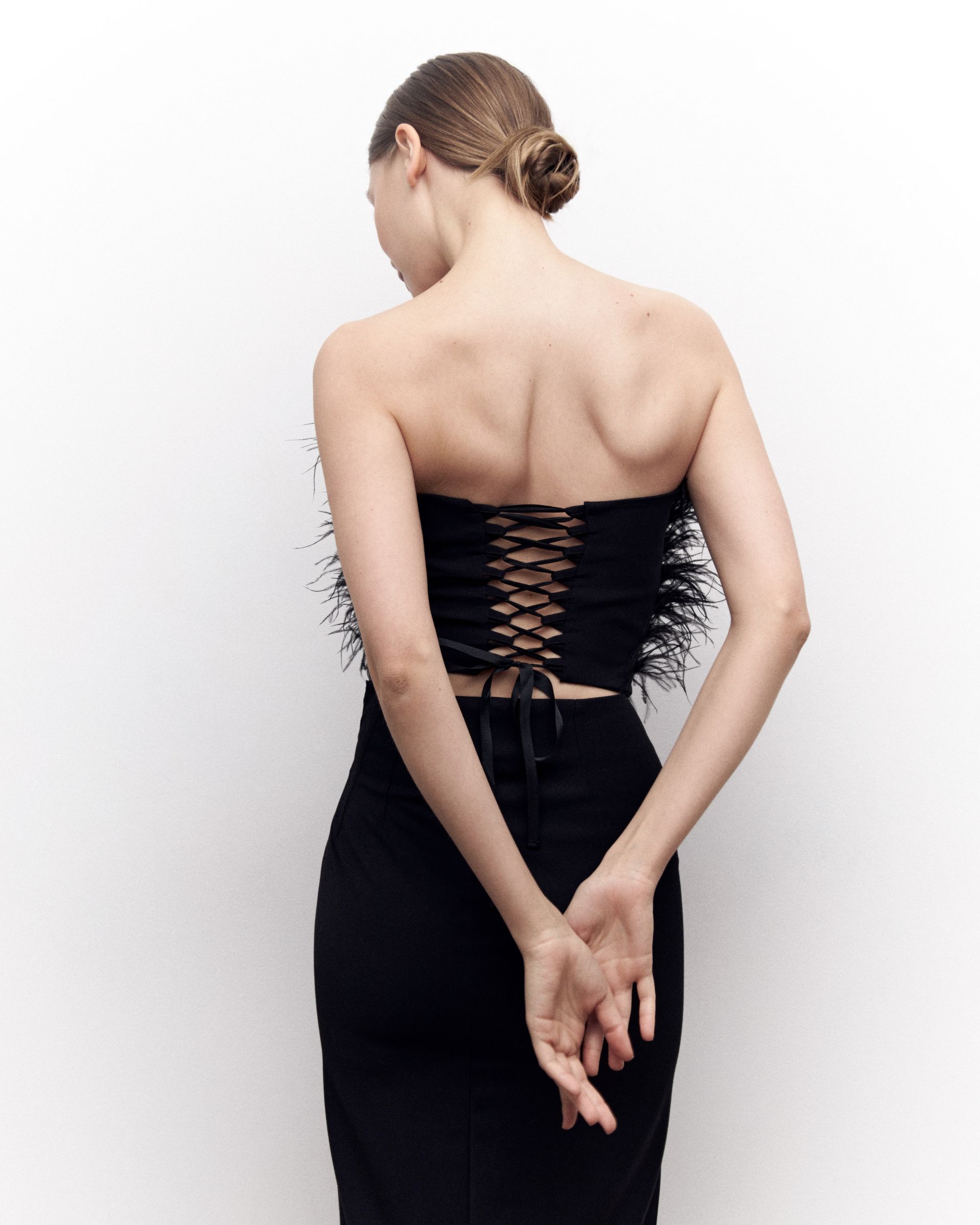 Woman in a black strapless corset and skirt, back to the camera, hands clasped behind back, against a white background.