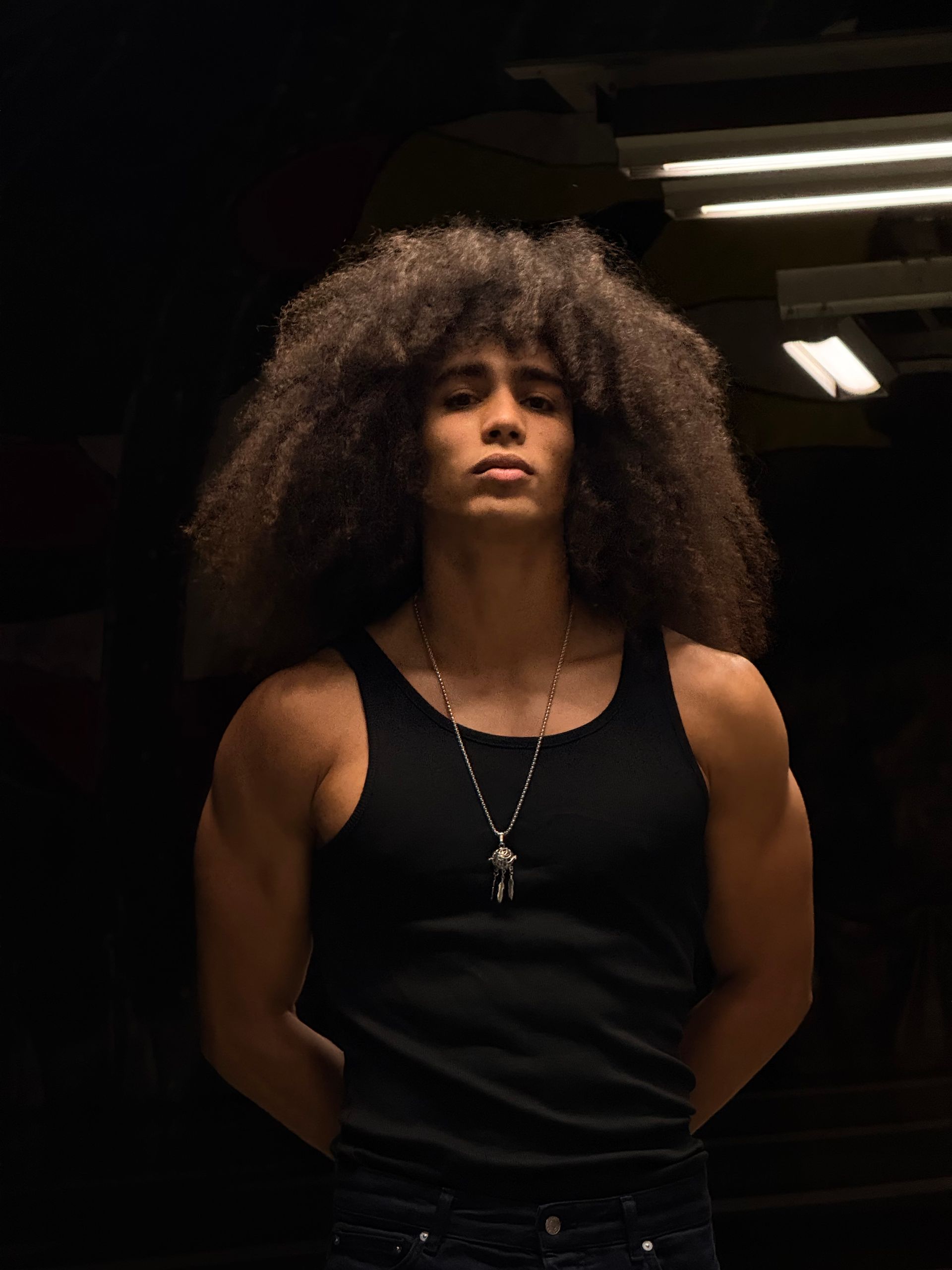 Man with large afro, arms crossed behind back, wearing black tank top. Standing in dim, dark area.