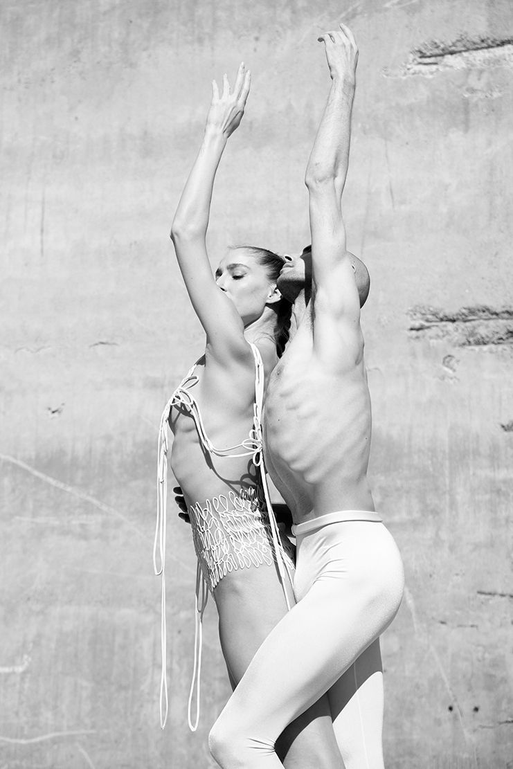 Two dancers reaching up, bodies entwined, against a textured wall; one in white, one in embellished attire.