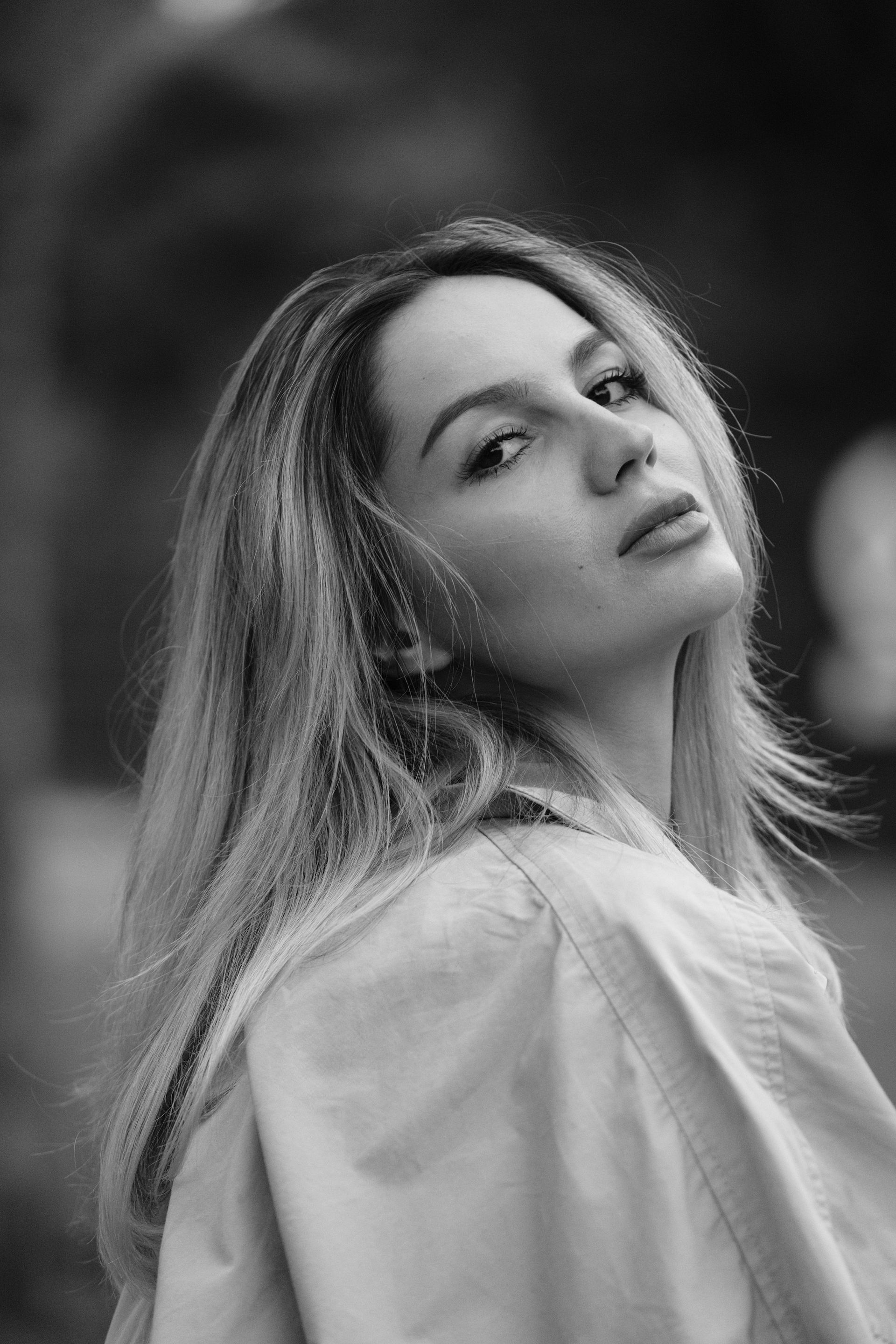 Woman with long blonde hair, looking over her shoulder, wearing a light-colored coat outdoors.