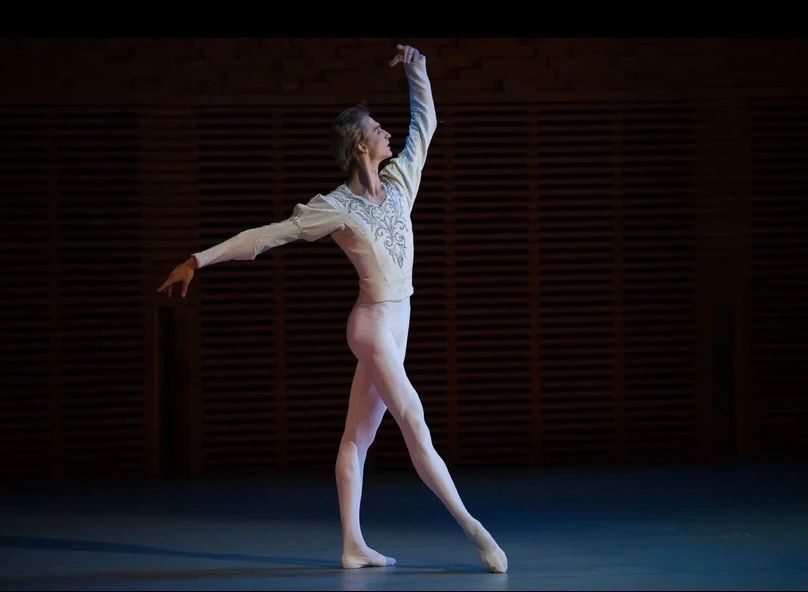 Male ballet dancer in a white and silver embroidered top and pink tights, poised on stage.