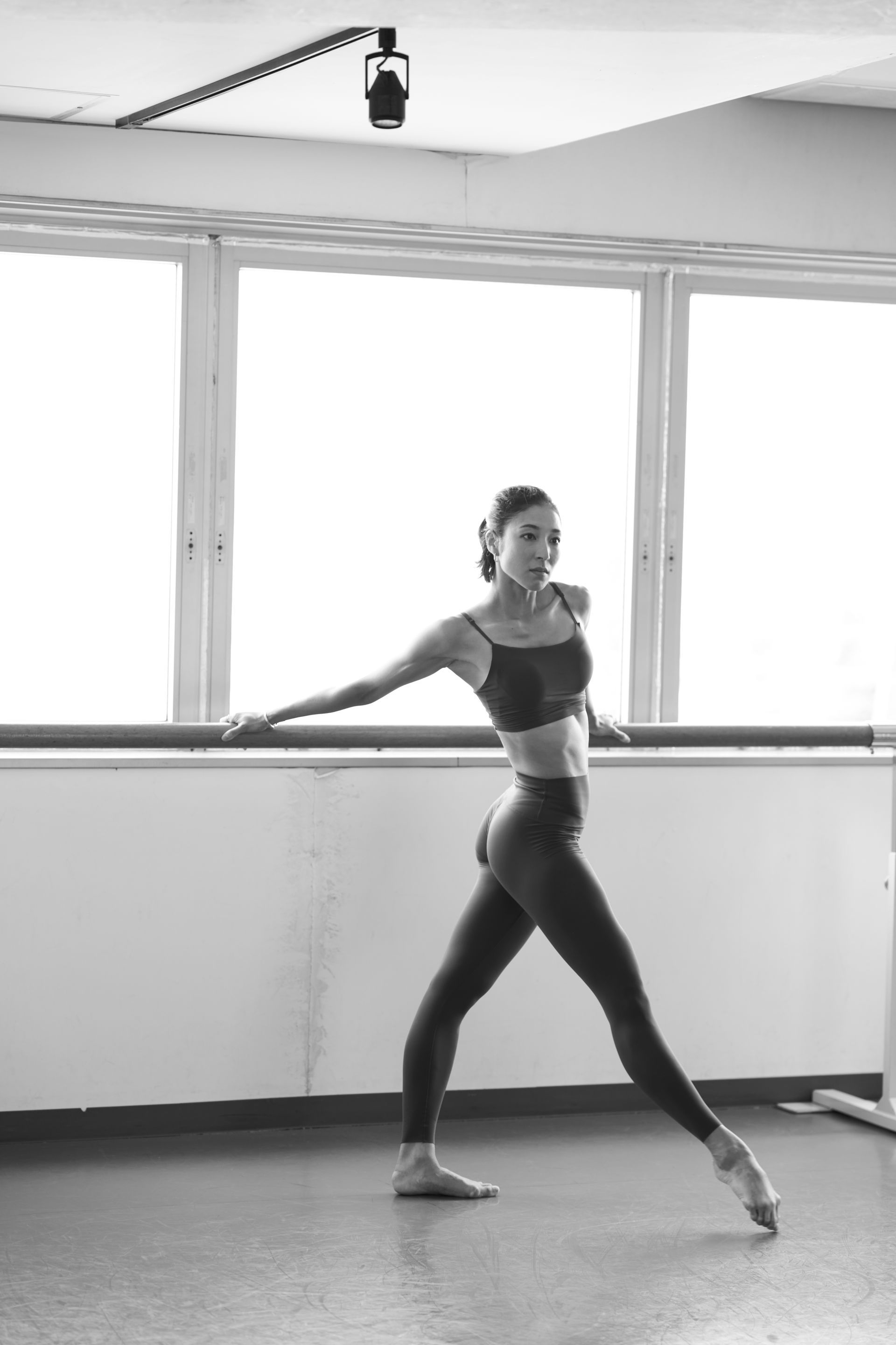 Ballet dancer in a studio, extending arm toward barre. Black and white photo.