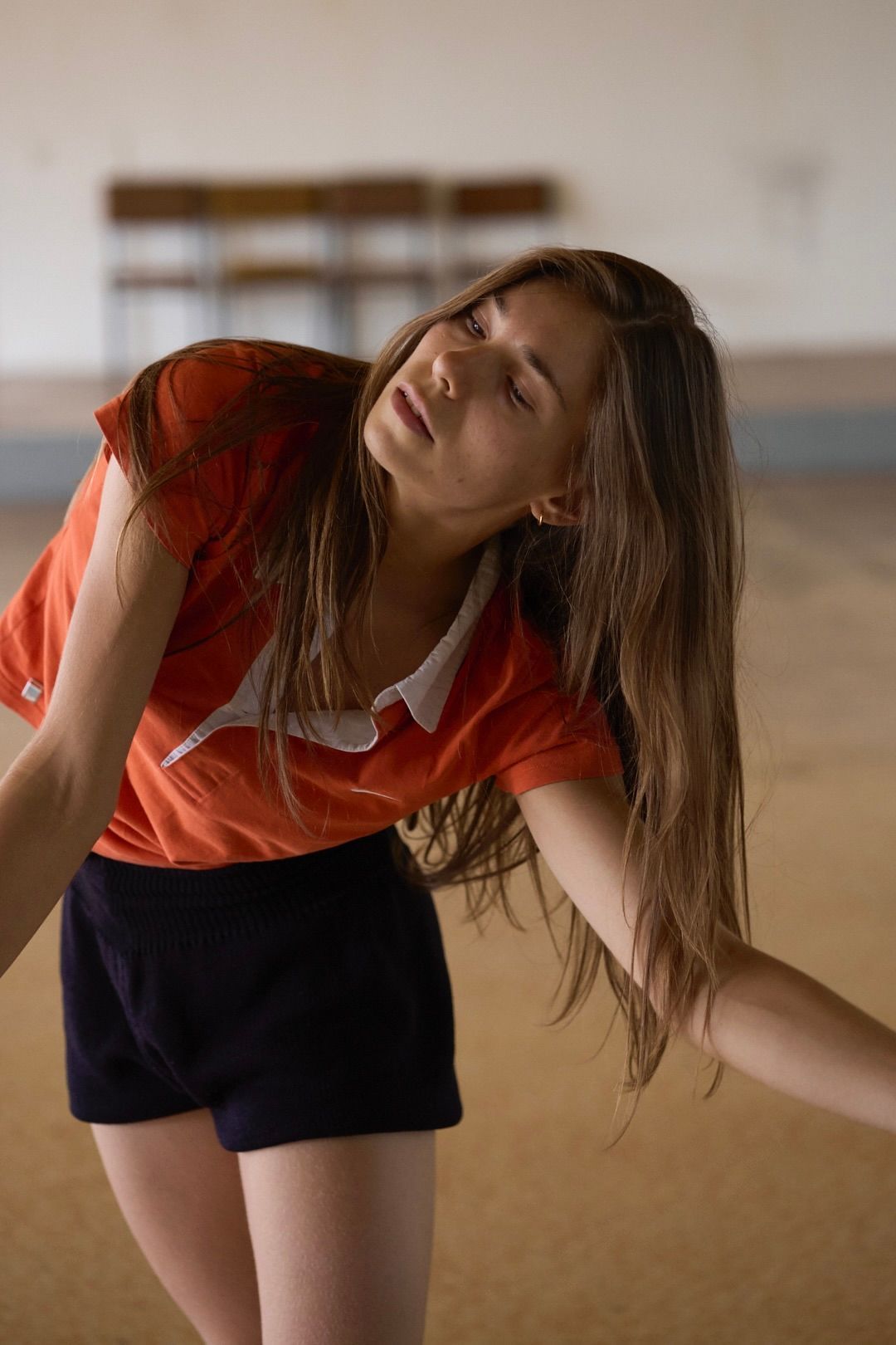 Woman in orange shirt and dark shorts, tilted head, eyes closed, arms outstretched in a gym or dance studio.