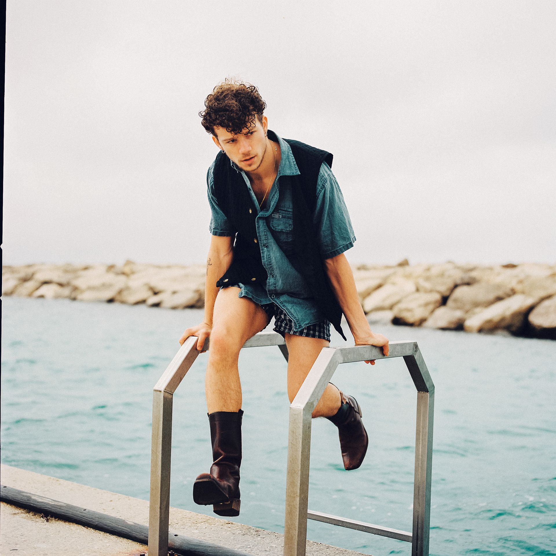 Man in denim shirt, vest, and boots sits on a metal rail, looking down at water.
