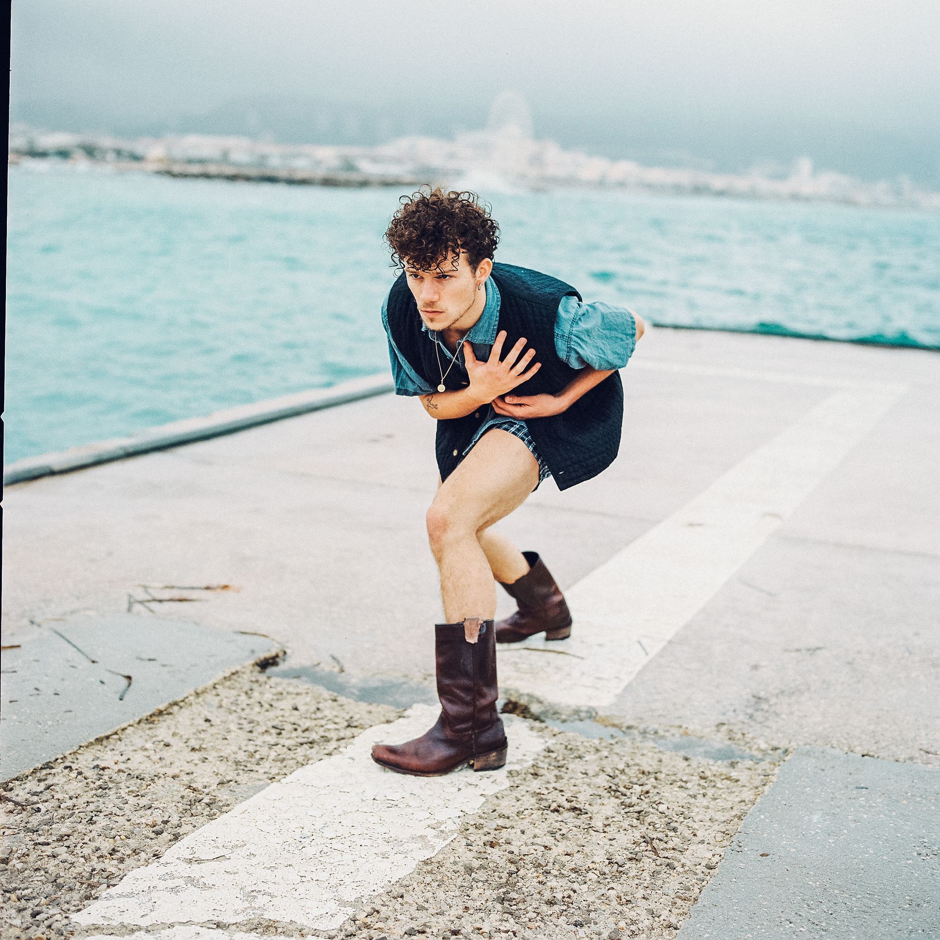 Man in boots, vest, and shorts, poses dramatically by the water, hands on chest.