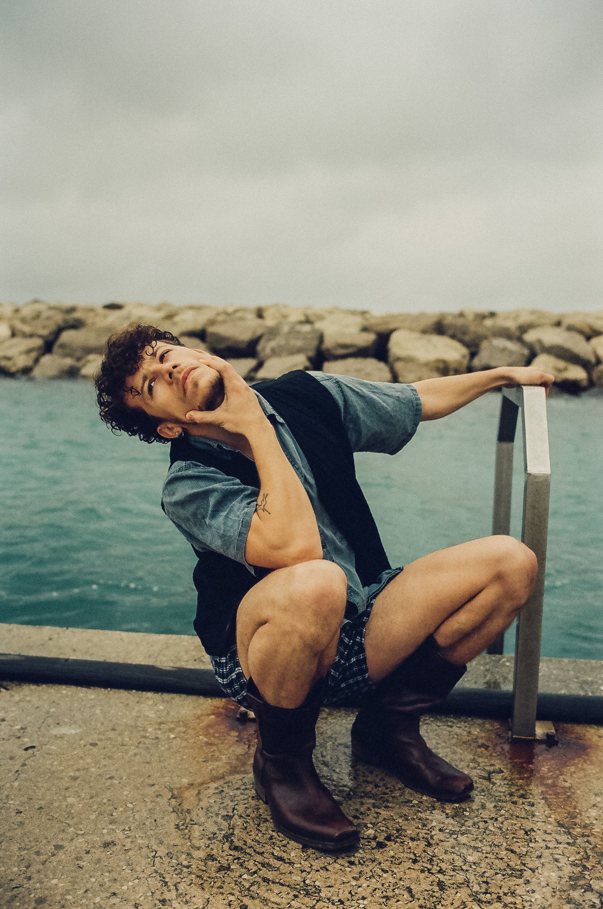 Man squatting, touching neck, by water. Wears vest, shorts, boots. Cloudy sky.