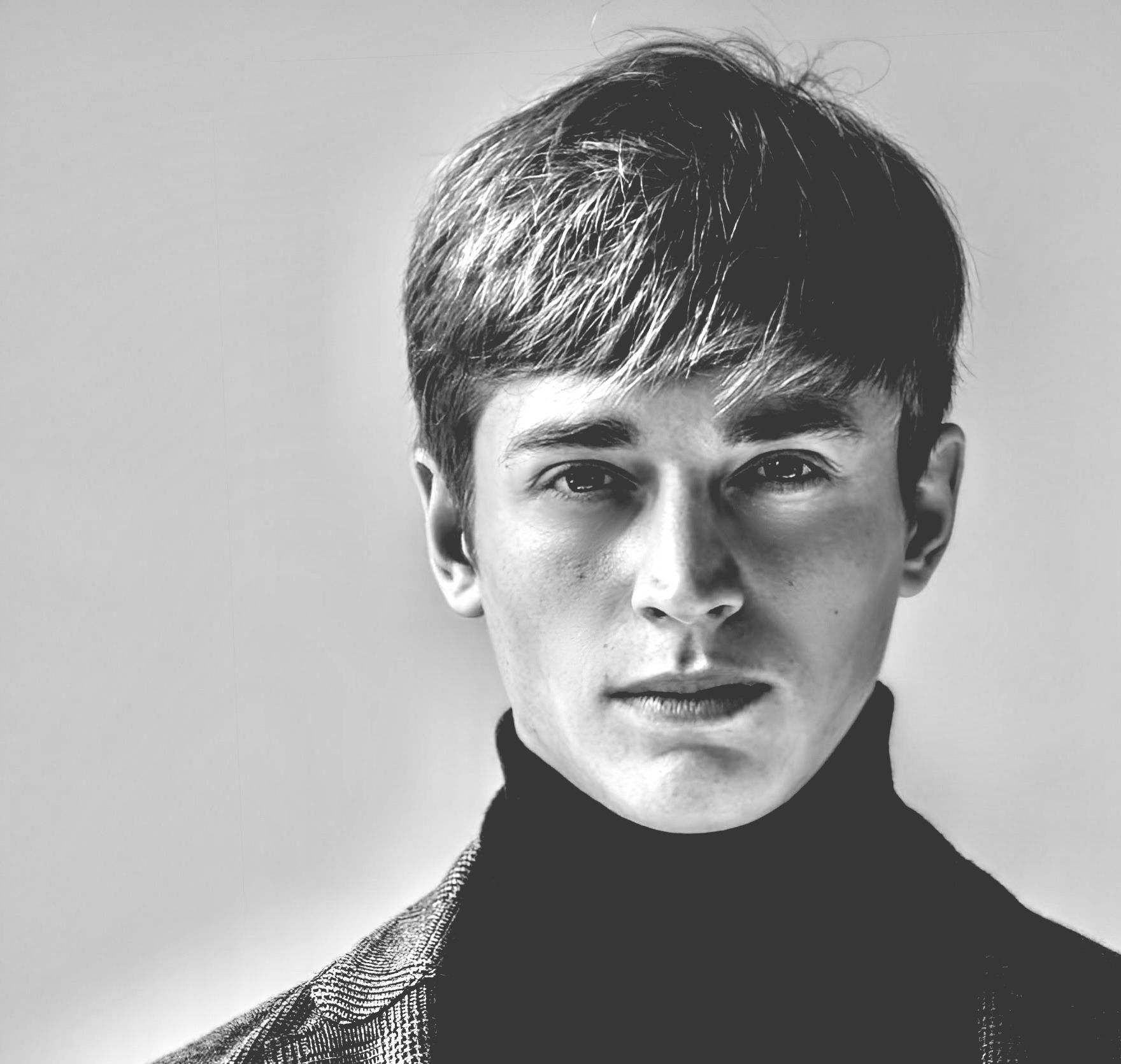 Man wearing a black turtleneck and patterned jacket, looking at the camera. Black and white.