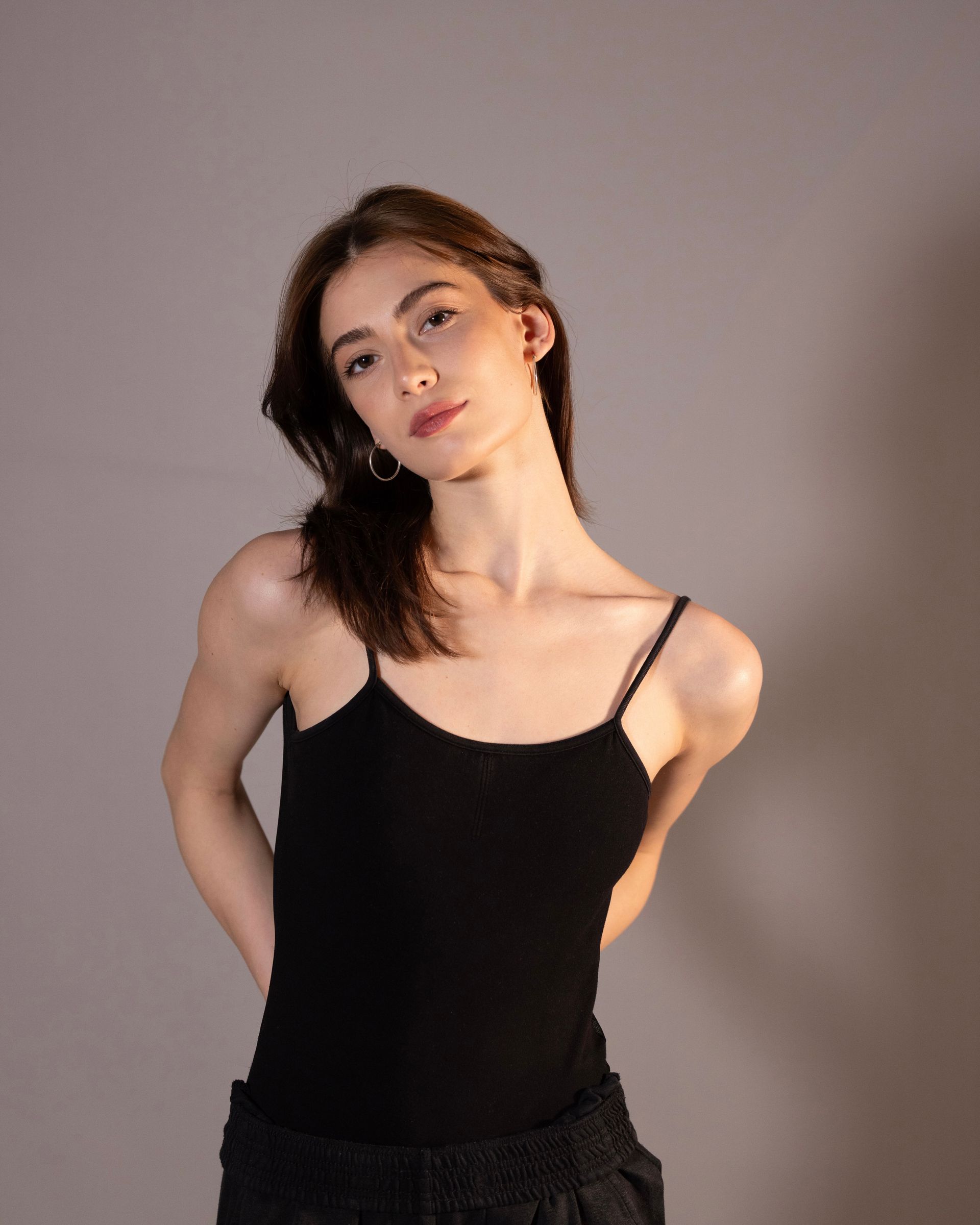 Woman in a black spaghetti strap top, looking at the camera, hands behind her back, against a grey background.