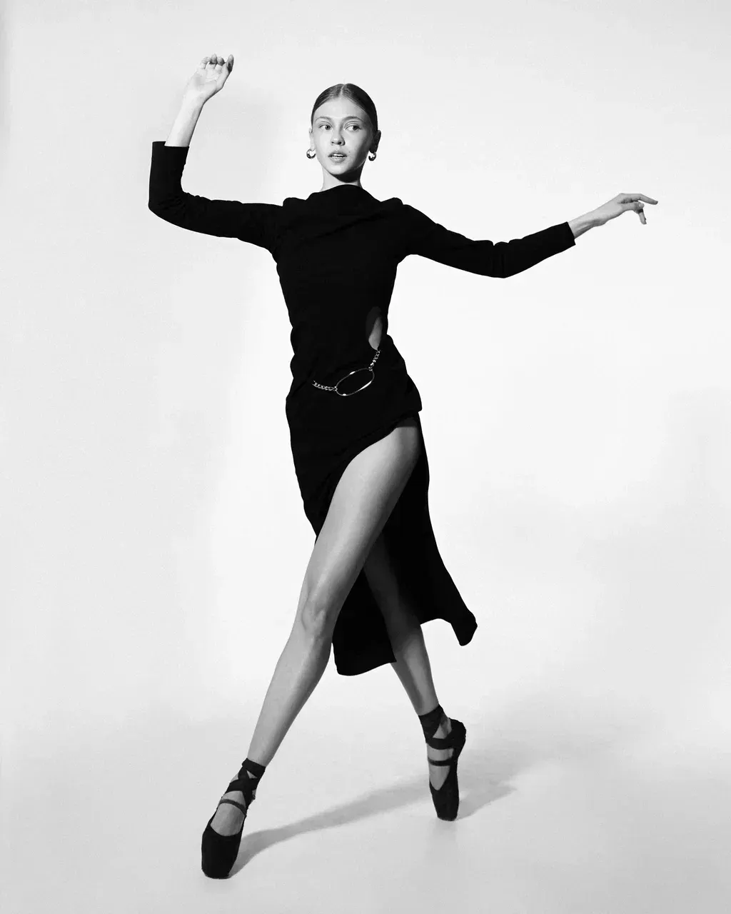 Woman in black dress and ballet shoes, in dance pose, raised arm, high slit.