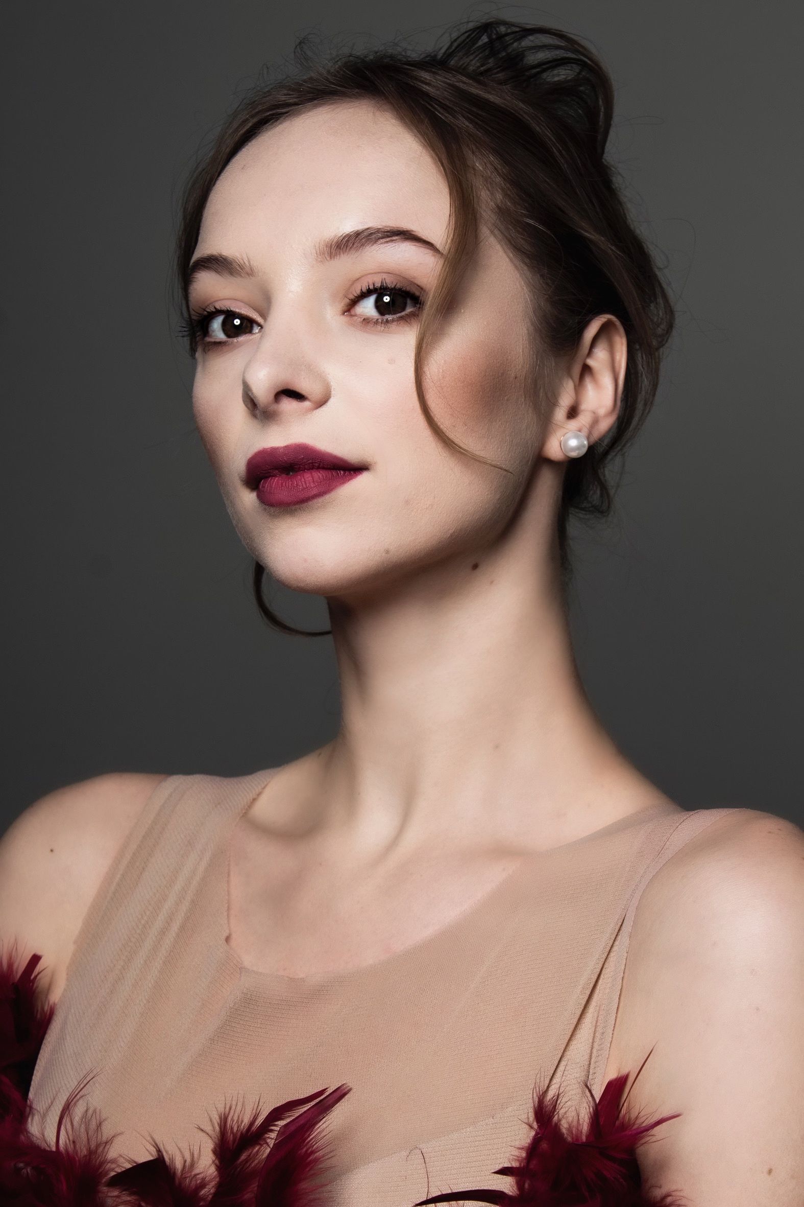 Woman with updo, dark red lipstick, wearing a beige sheer top with red feathers, against a gray backdrop.