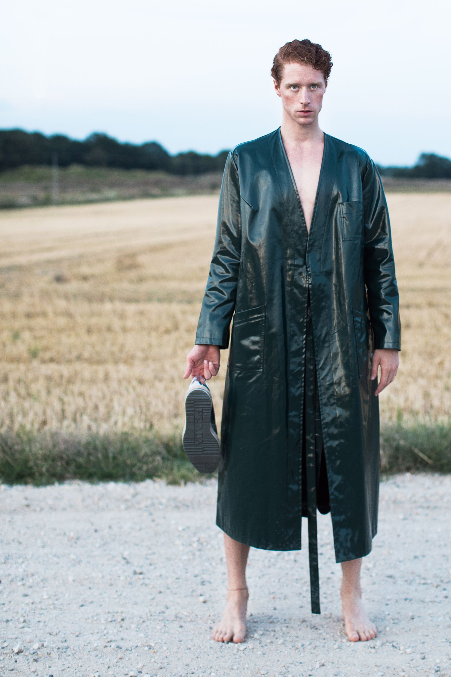 Man in green coat, barefoot, holding shoe, standing on dirt road, field background.