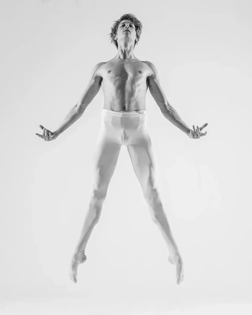Ballet dancer leaps, arms outstretched, wearing white tights and pointe shoes.