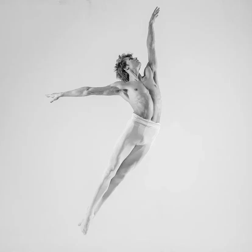 Male ballet dancer leaps gracefully, arms outstretched, studio setting, black and white.