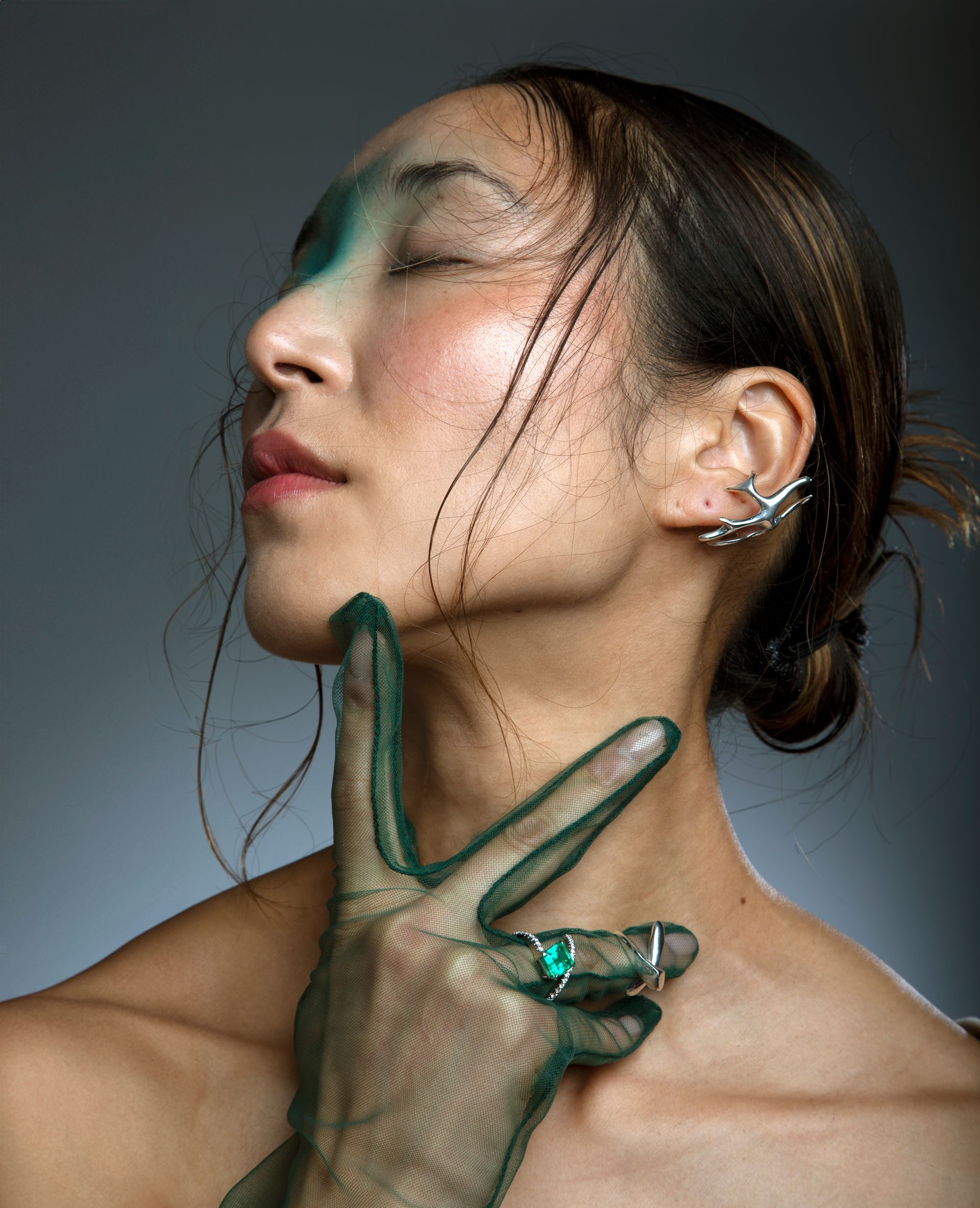 Woman with eyes closed, hand in green netting at chin. Silver ear cuff and ring. Green and pink makeup, wet hair.
