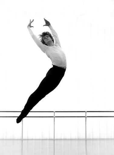 Ballet dancer leaps in the air, arms raised, black pants, white shirt, studio background.