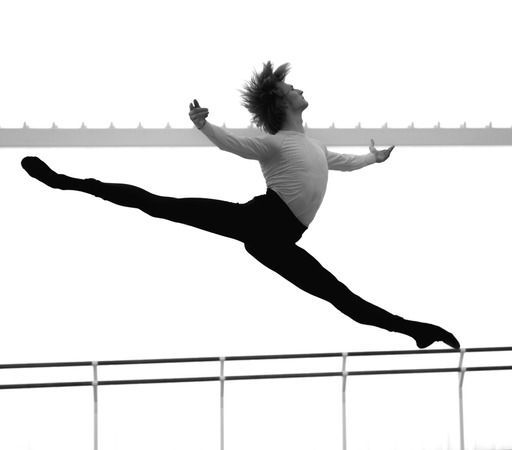 Male ballet dancer leaps in a split, arms outstretched. Black and white photo against a bright sky.