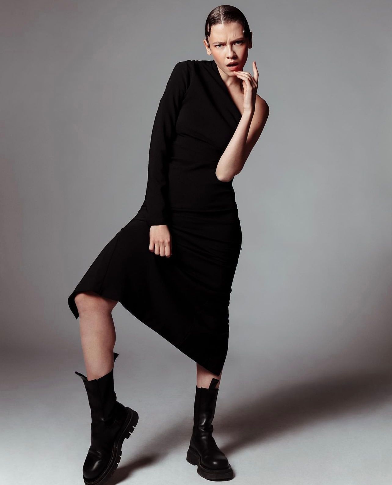 Woman in black dress and boots, posing with hand near face. Gray background.