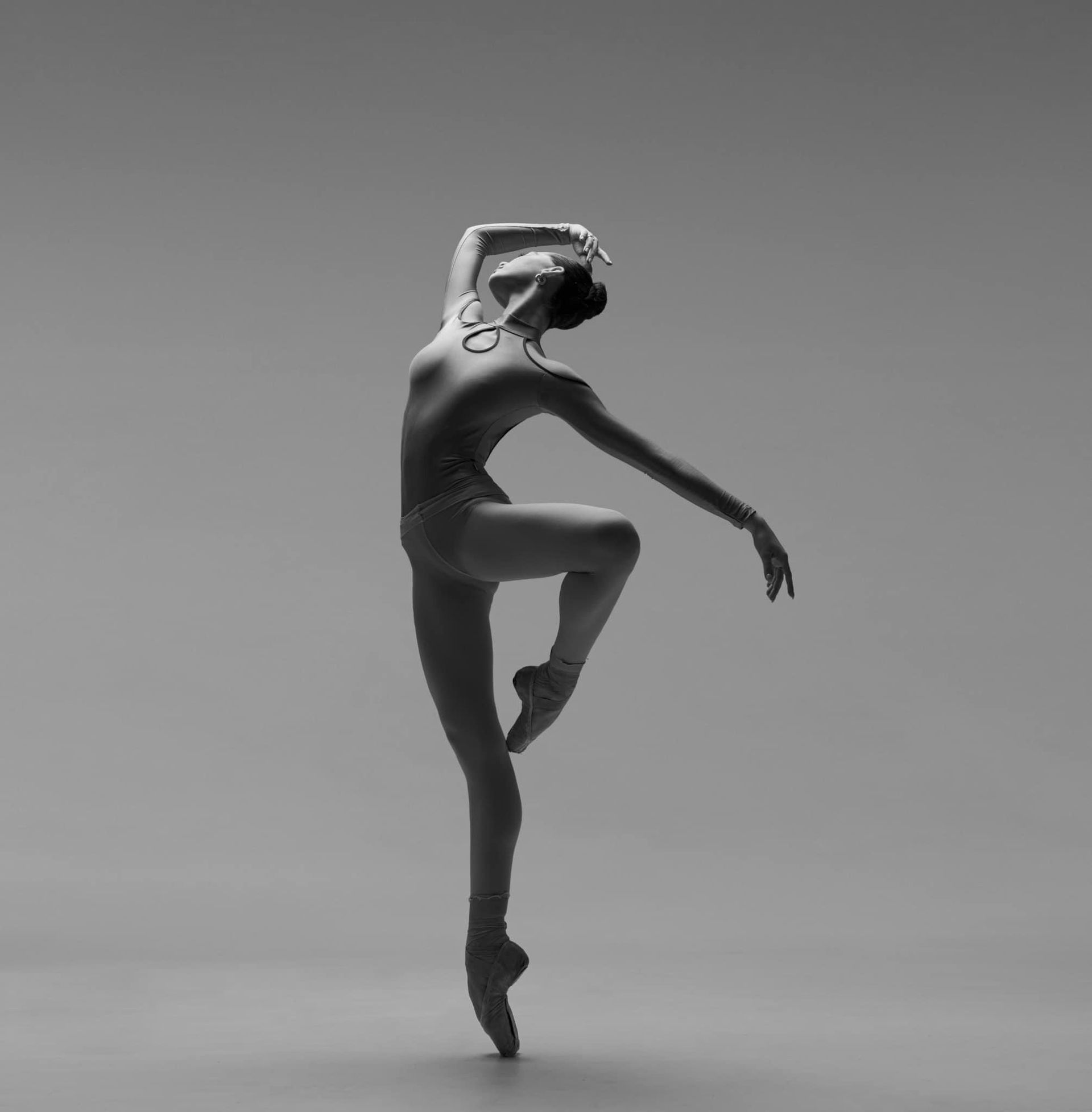Ballerina en pointe, arms gracefully extended, body arched against a gray backdrop.