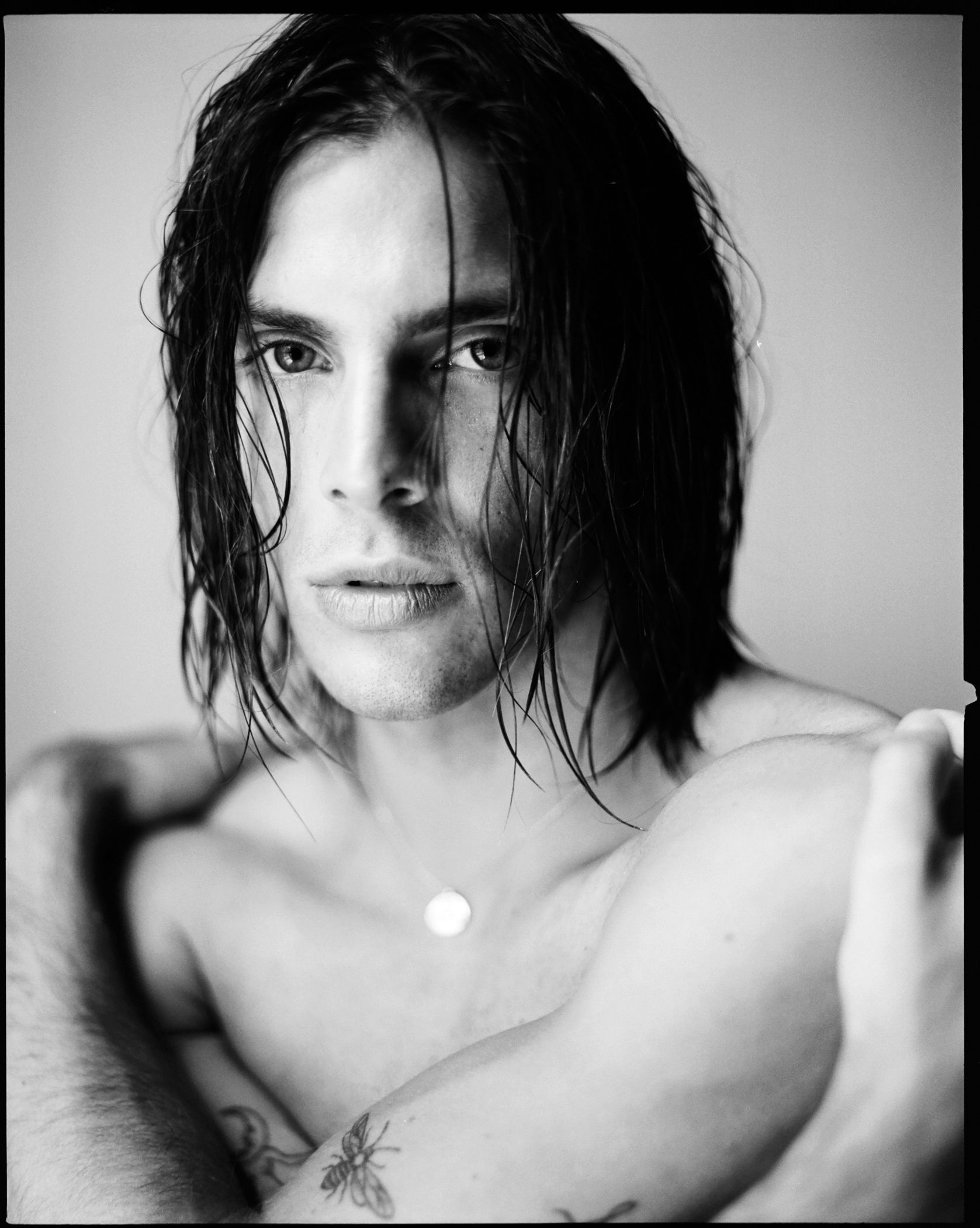 Man with wet, dark hair, looking intently, arms crossed, small necklace, tattoo of insect on arm.