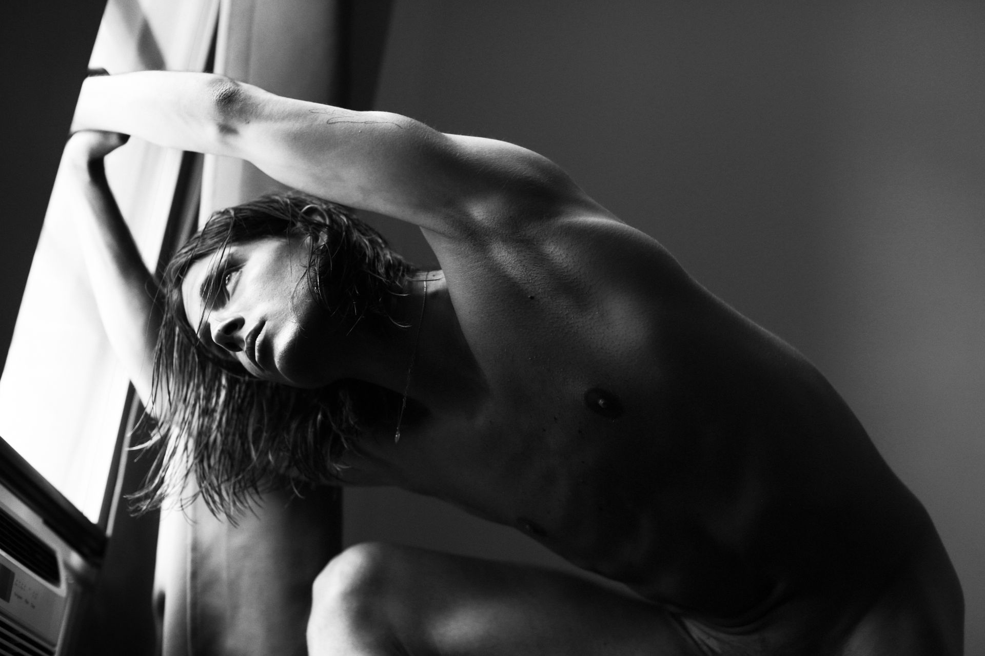 Nude person, in silhouette, bending near a window with sunlight.