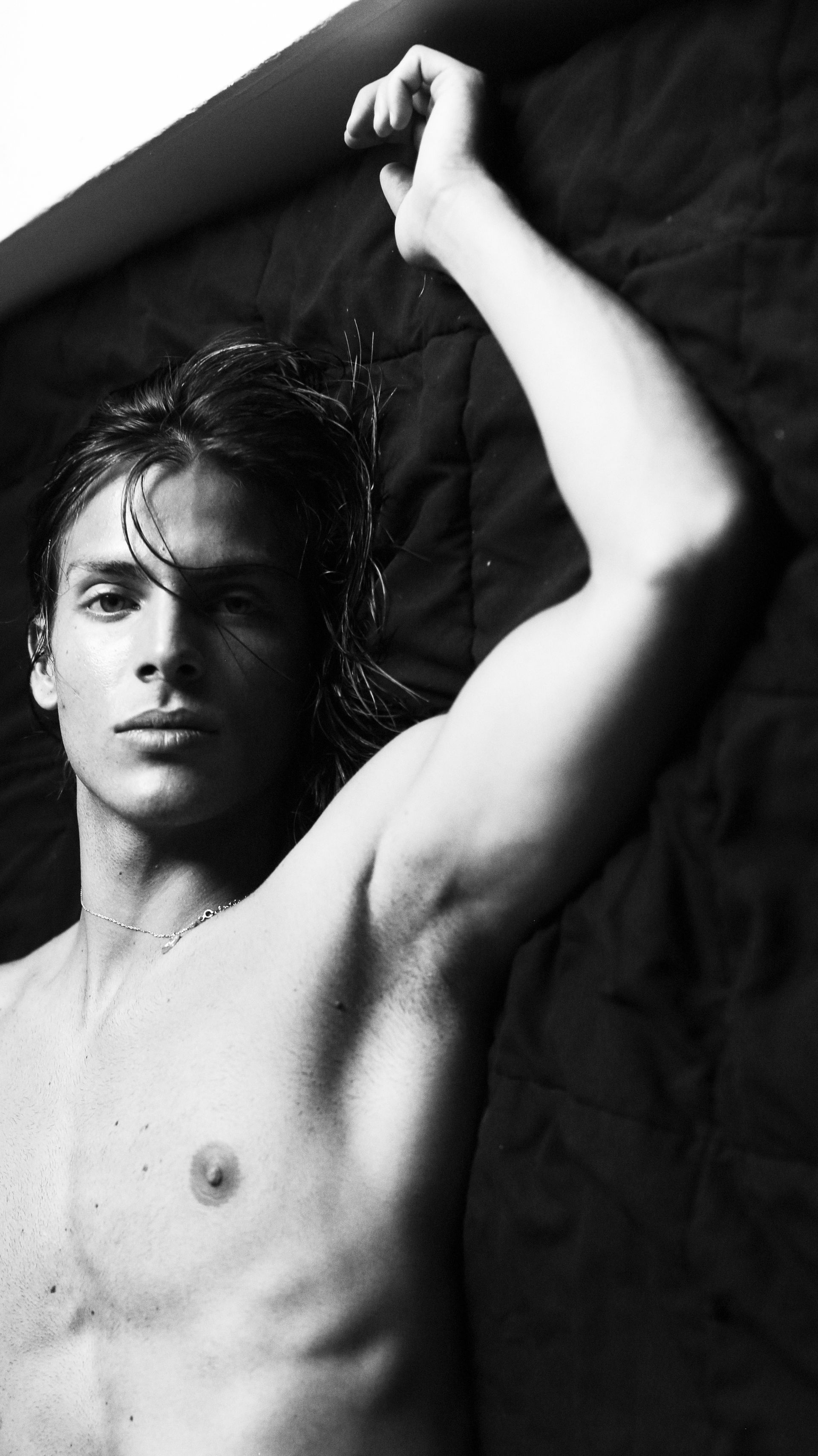 Man lying shirtless on dark surface, arm raised above head, looking at the camera. Black and white.