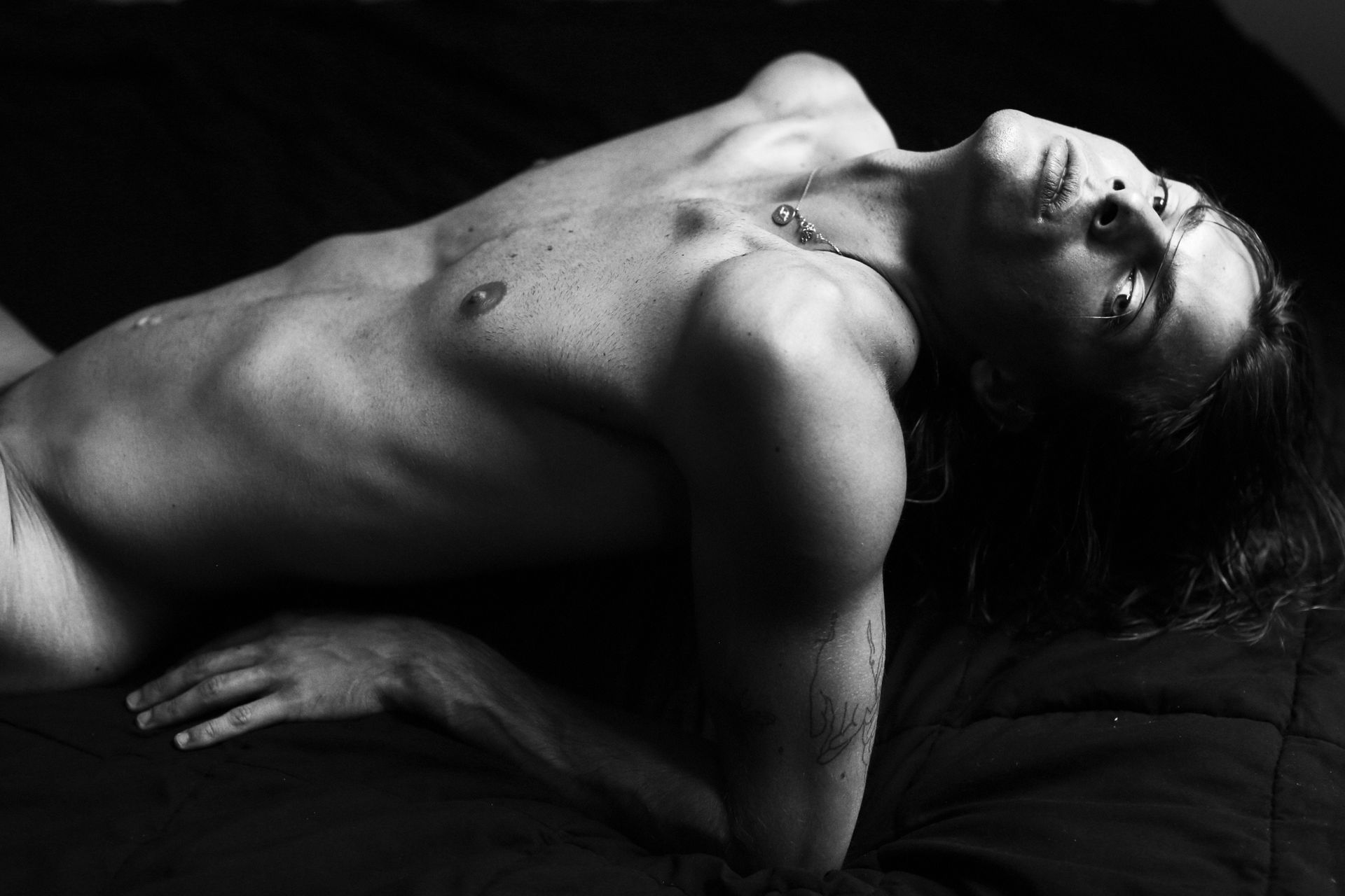 Man reclining, eyes closed, head tilted back, torso bare, wearing a necklace, in shadow.