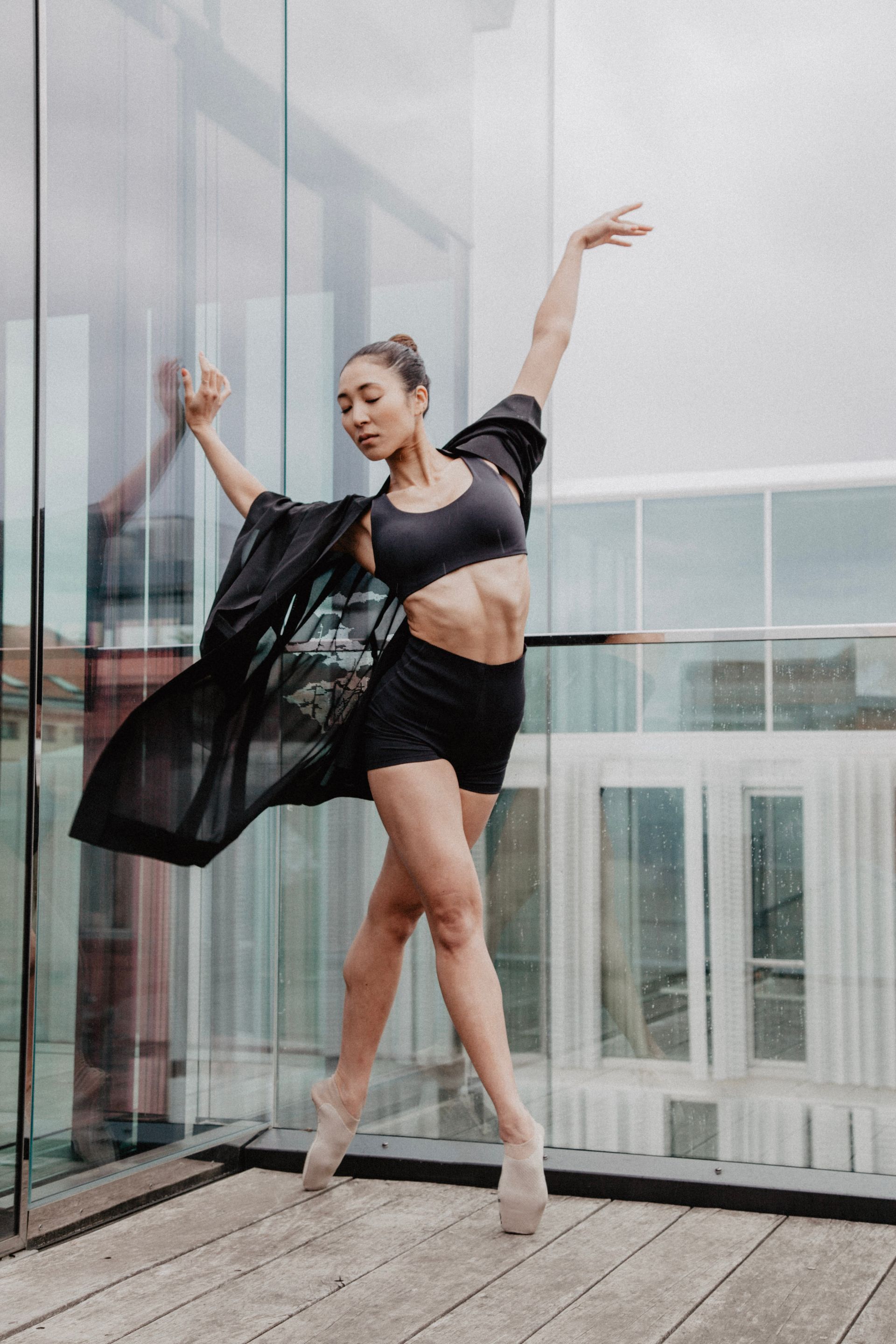 Ballet dancer in black outfit, en pointe, arms raised on a wooden deck with glass windows.
