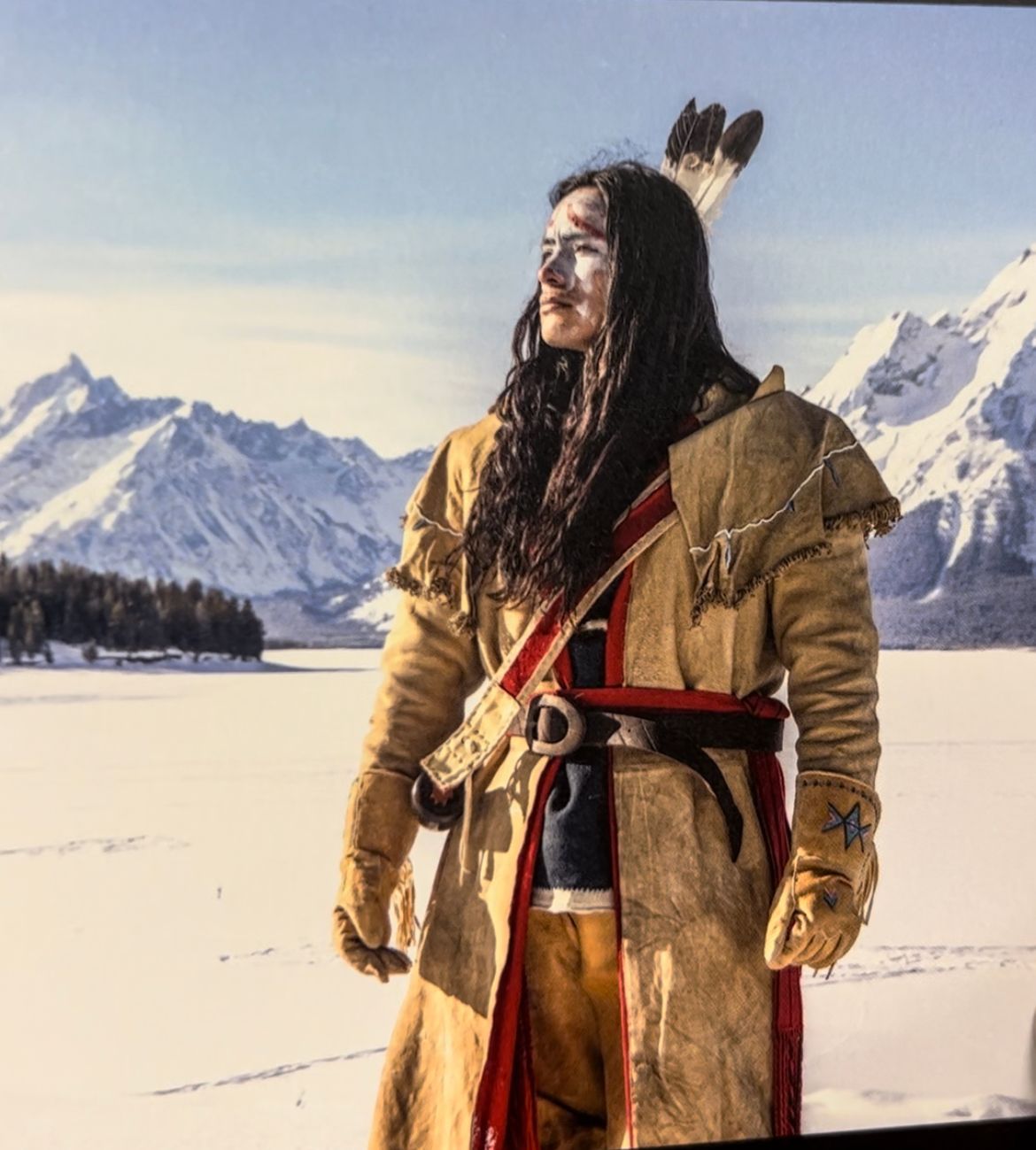 Man in buckskin coat and feather headdress, standing by a snowy lake with mountains in background.