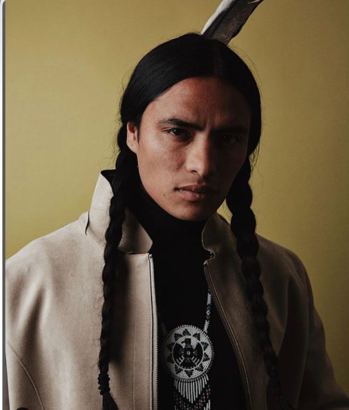 Man with long braids and feather in hair, wearing a beige jacket and a black turtleneck, looking at the viewer.