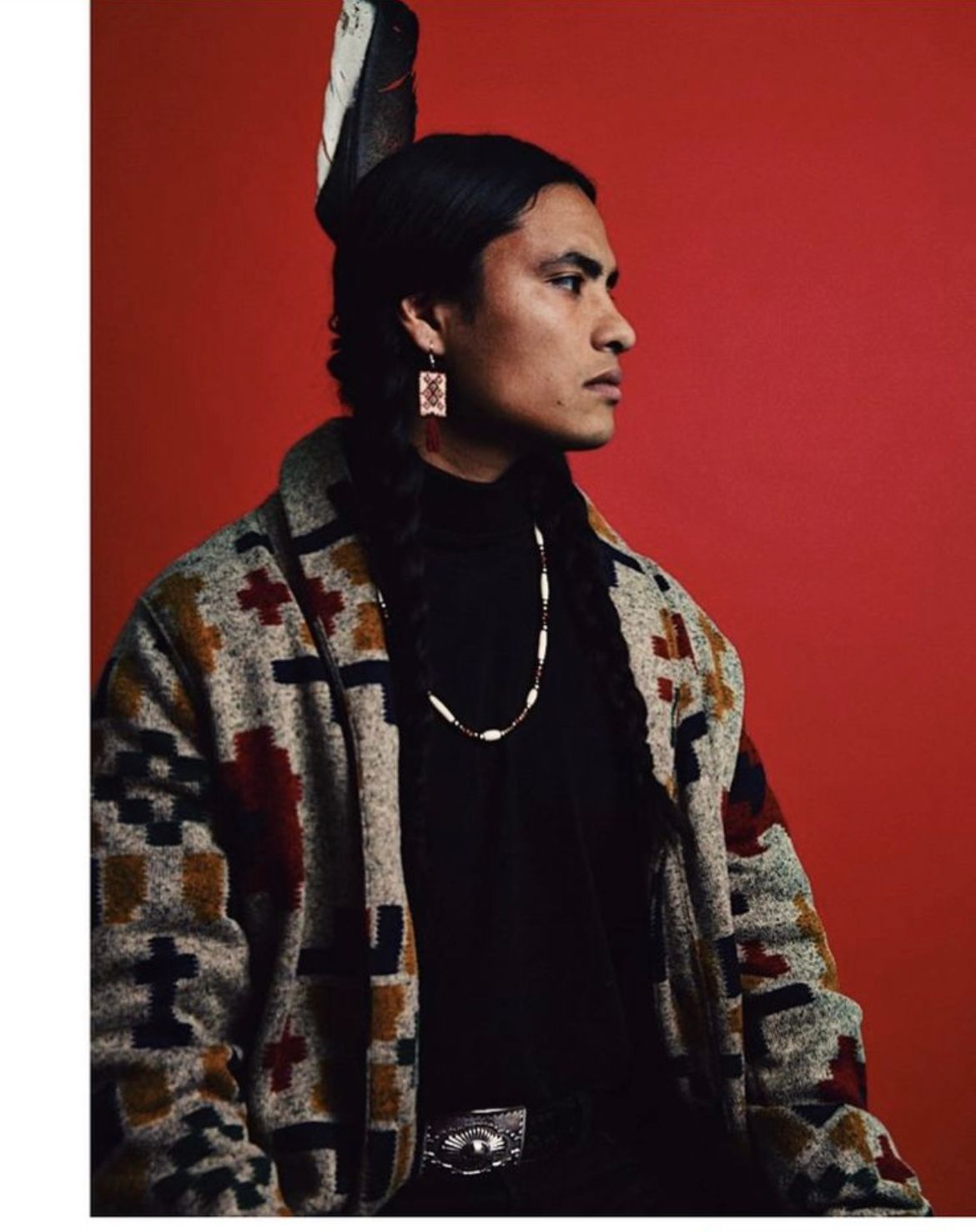 Person in patterned jacket, with feather headdress and jewelry, against red background.