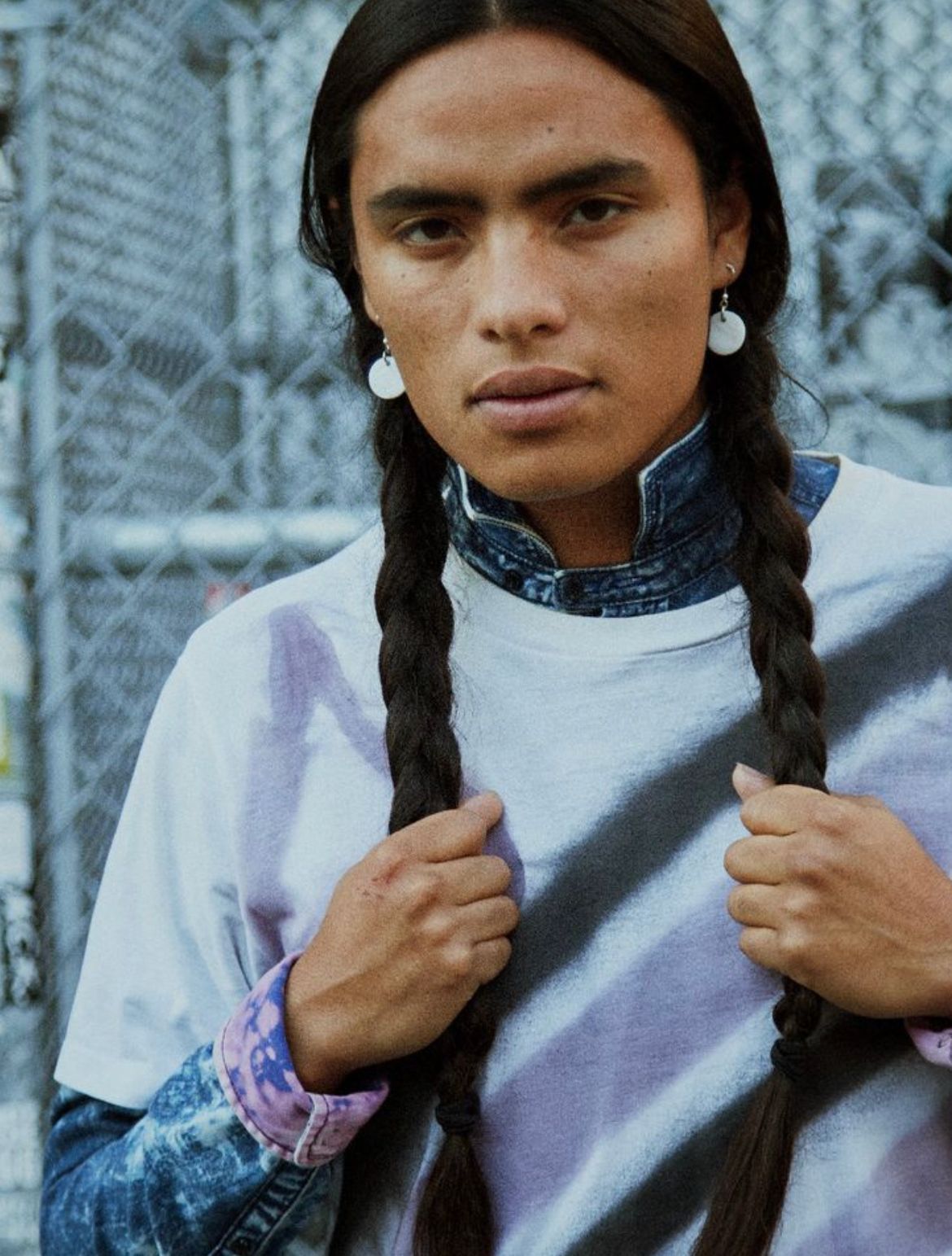 Person with long braids holds them, wearing white t-shirt and earrings. Gray and purple tie-dye, denim collar.