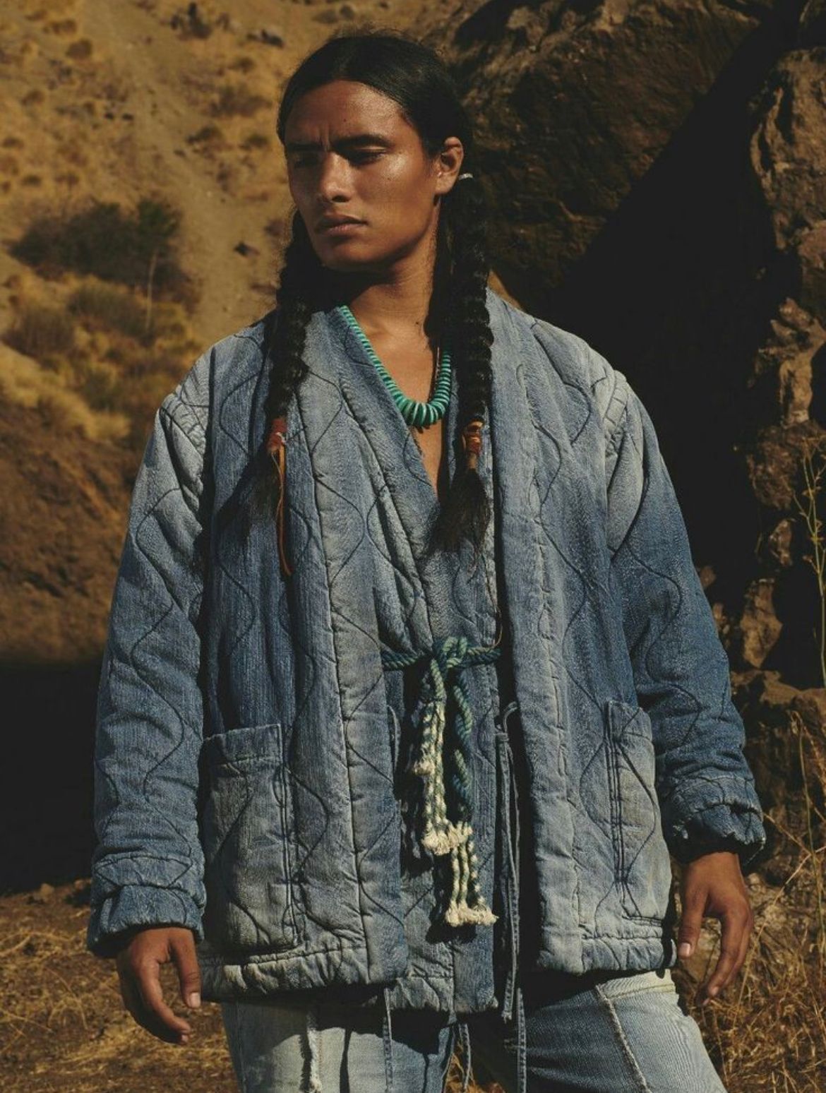 Person in blue quilted jacket with braids, turquoise necklace, and denim pants, standing outdoors near rocks.