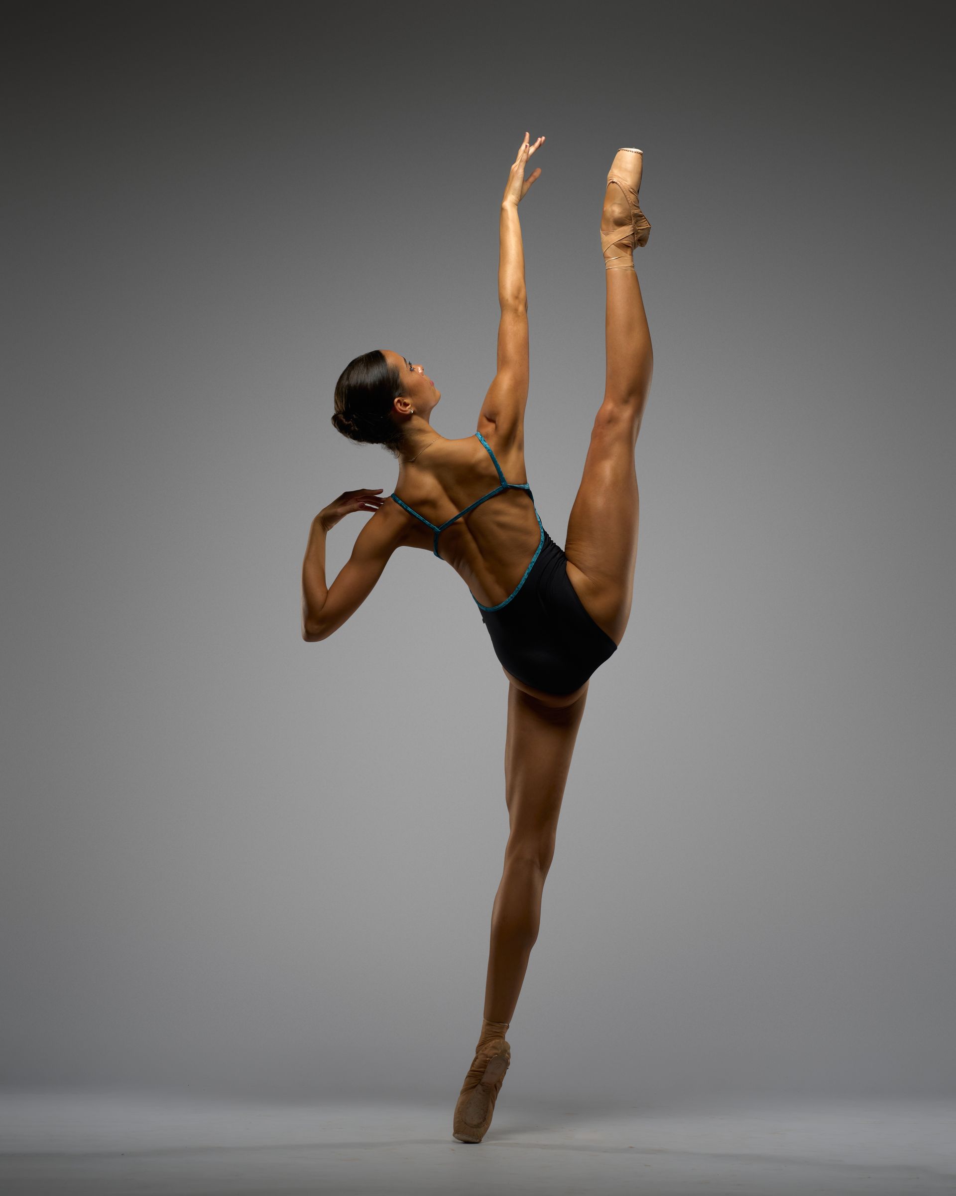 Ballerina in black leotard, balancing in a high split, arm raised overhead, wearing pointe shoes, studio setting.