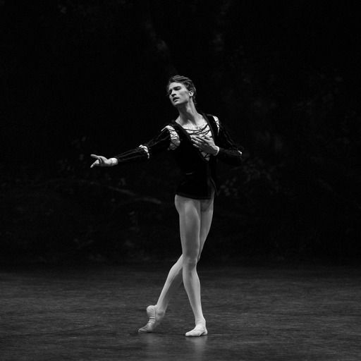 Male ballet dancer performing on stage, wearing a black top, tights, and a focused expression.