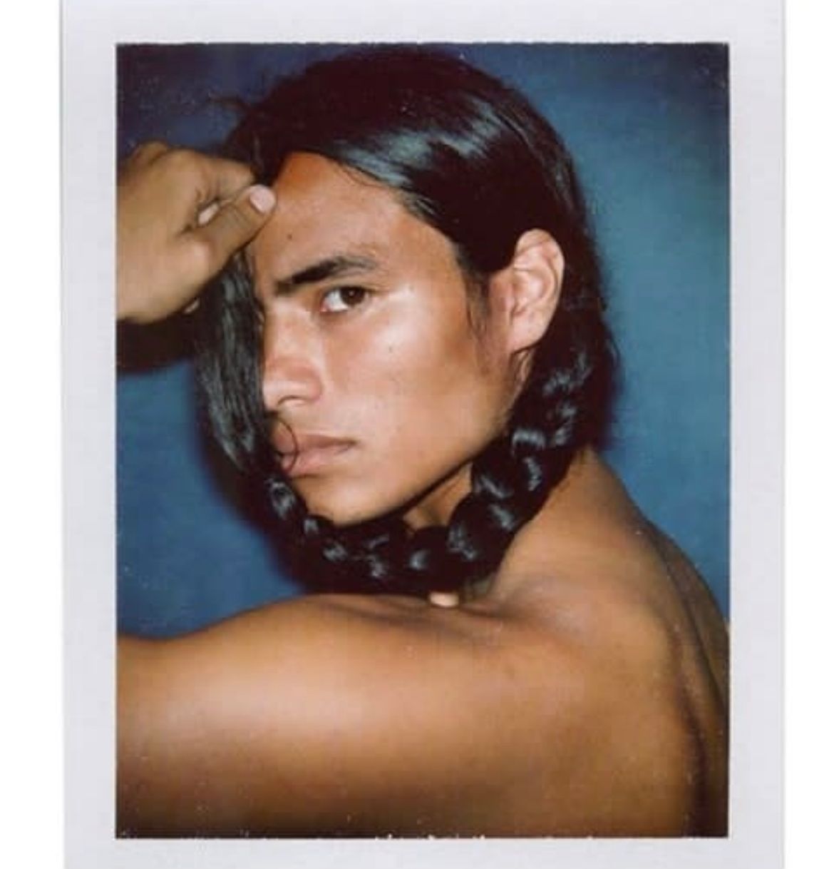 Person with long dark hair in a braid, posing. Bare chest, blue background, serious expression.