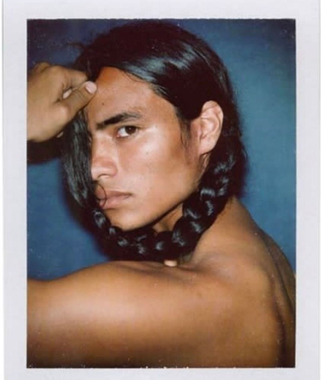 Man with dark hair in a braid, arm raised, looking at the camera.