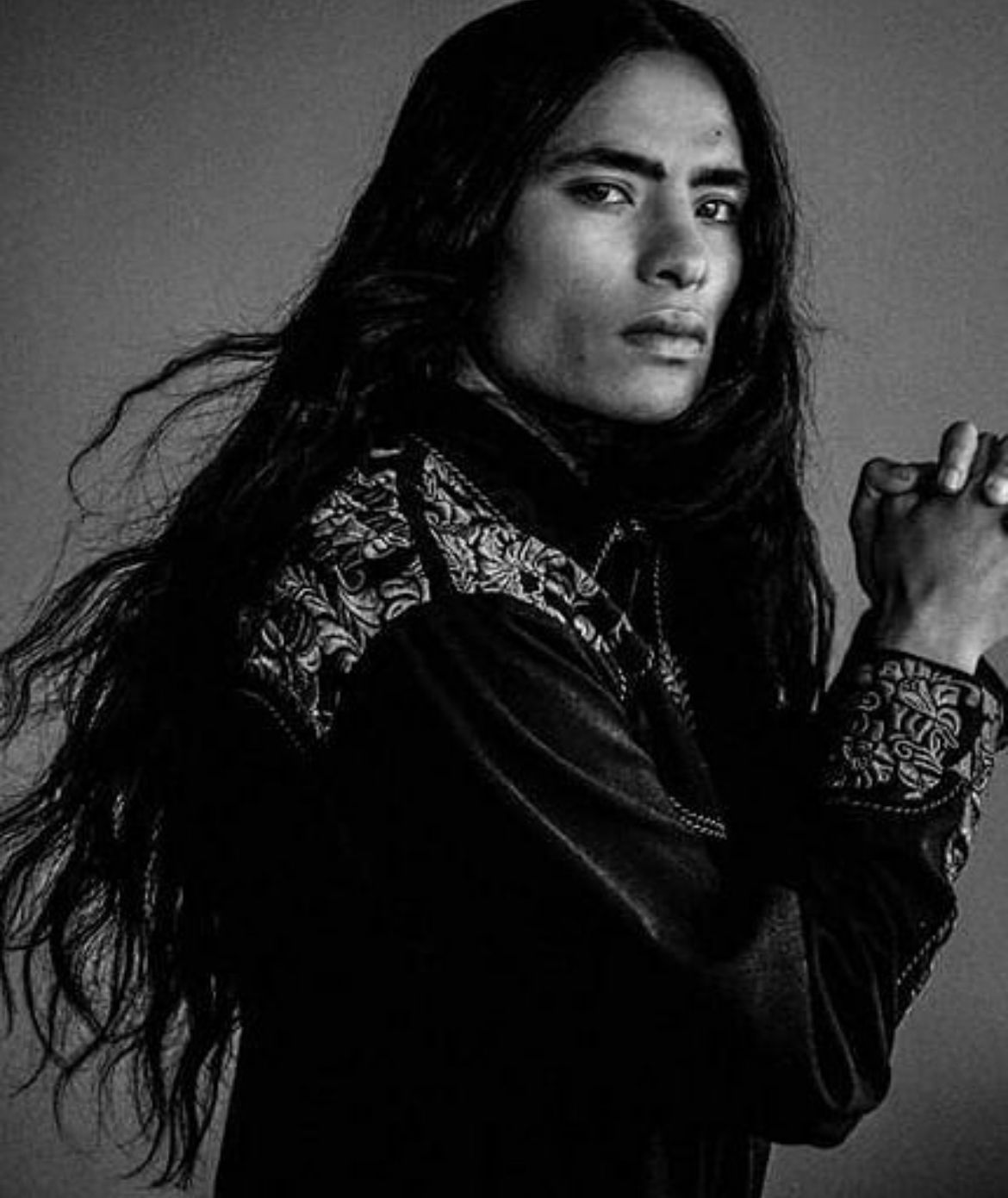 Man with long dark hair, wearing a dark shirt with embroidered detailing, hands clasped, looking at camera.
