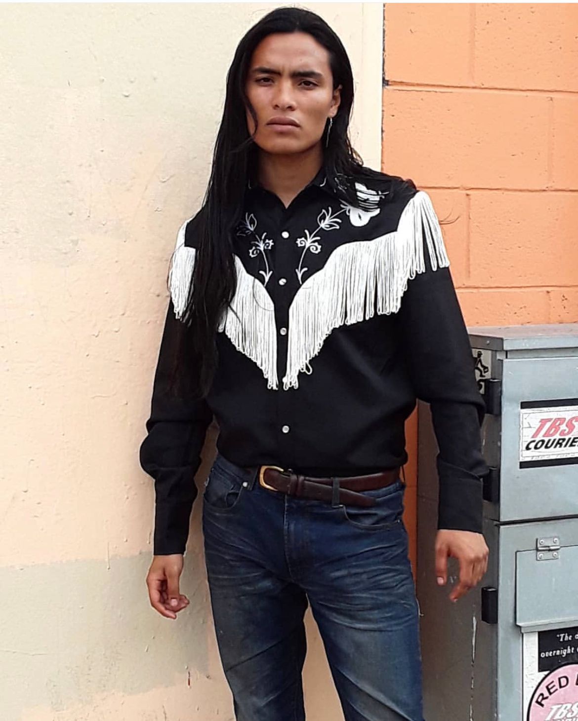 Man with long dark hair, wearing a black shirt with fringe and jeans, standing by a wall.