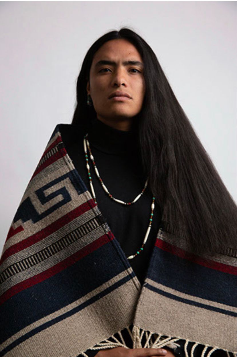 Person with long dark hair, wearing a black turtleneck and patterned blanket. They look forward.