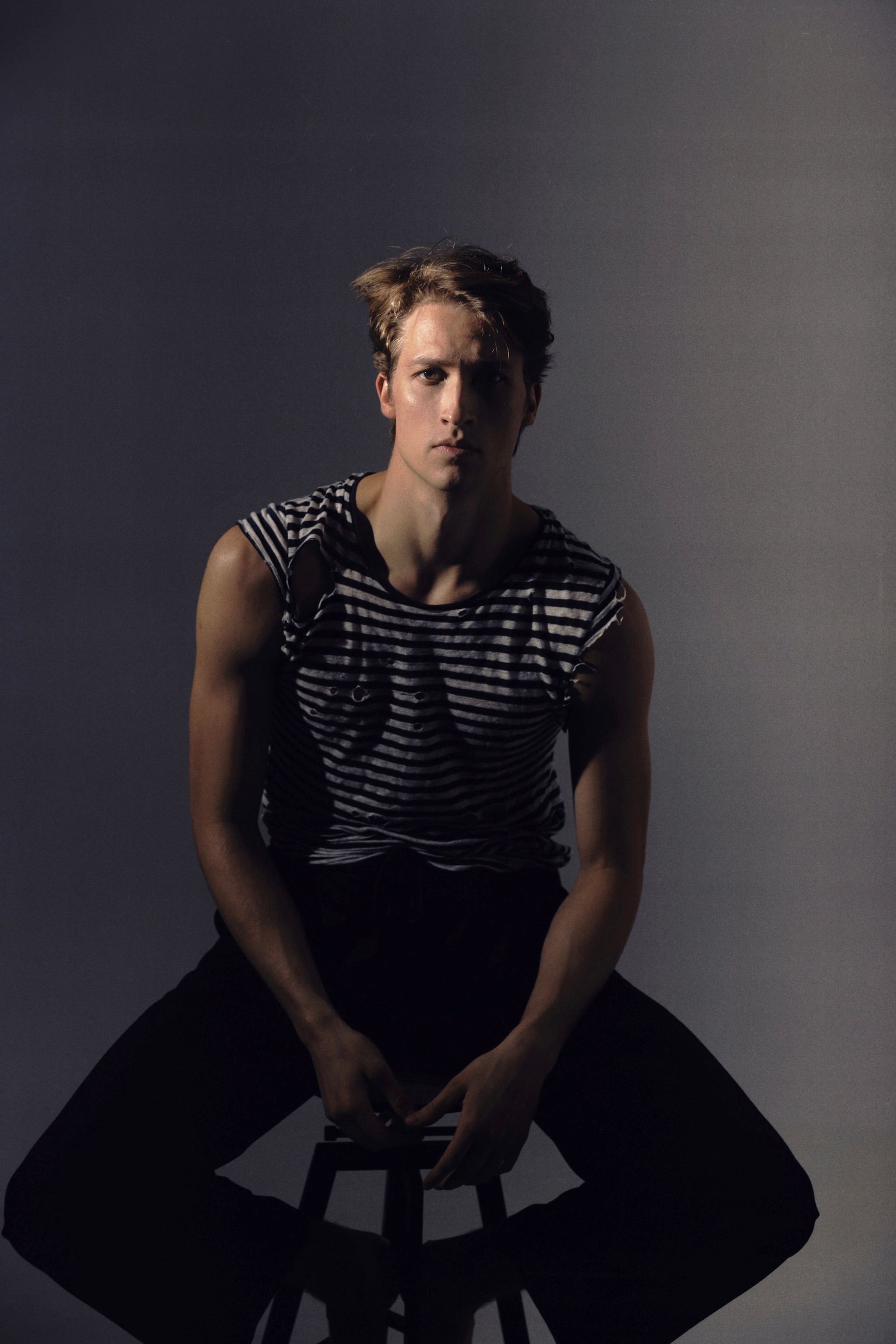 Man seated, wearing striped top and dark pants, gazing forward. Lit by spotlight on gray background.