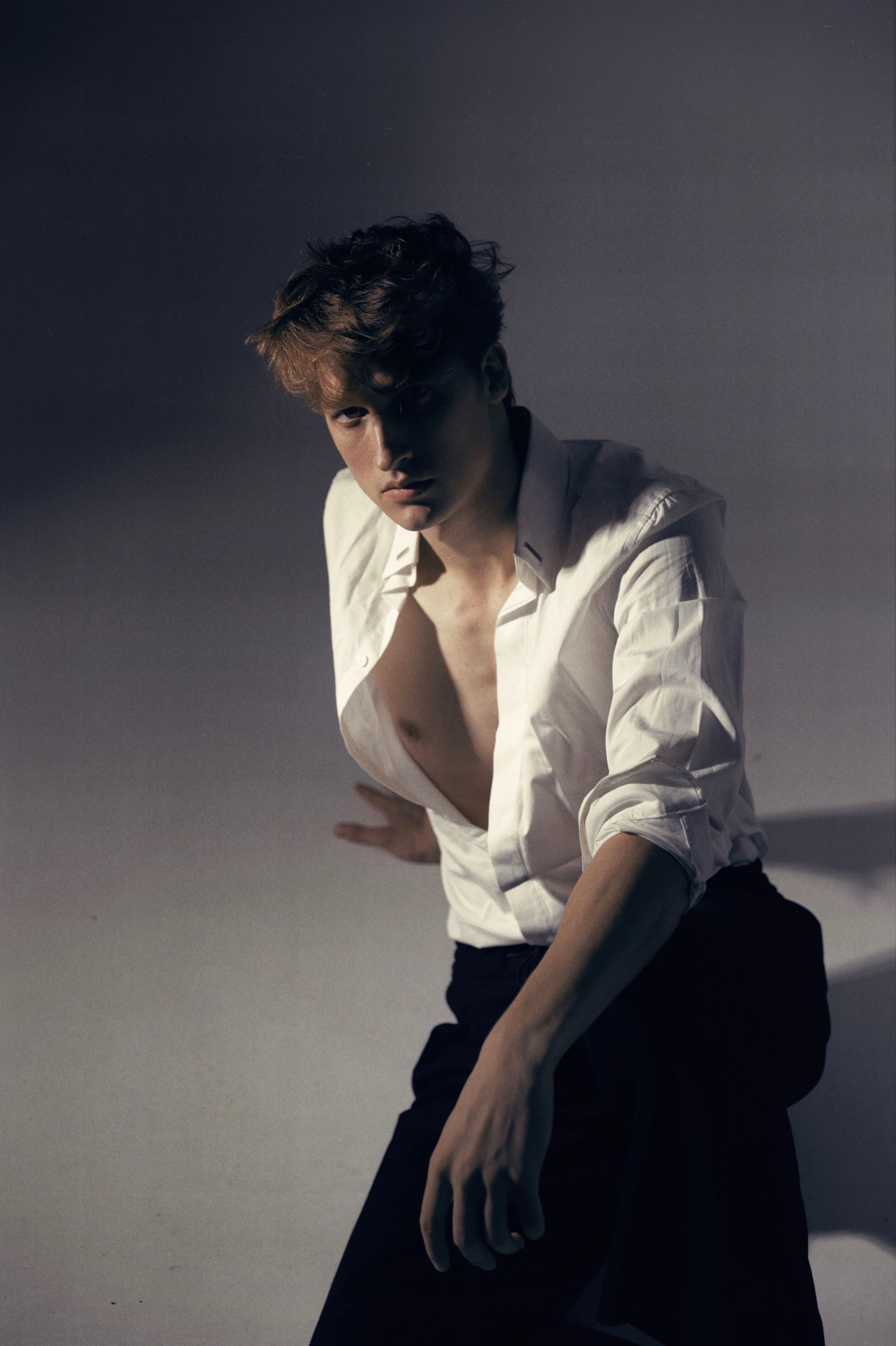 Man in white shirt, dark pants. Leaning, shirt open. Dramatic lighting, gray background.