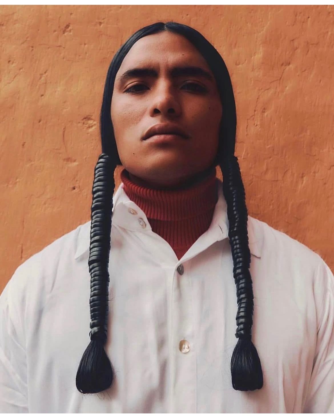 Person with long braids, wearing a white shirt and red turtleneck, in front of an orange wall.
