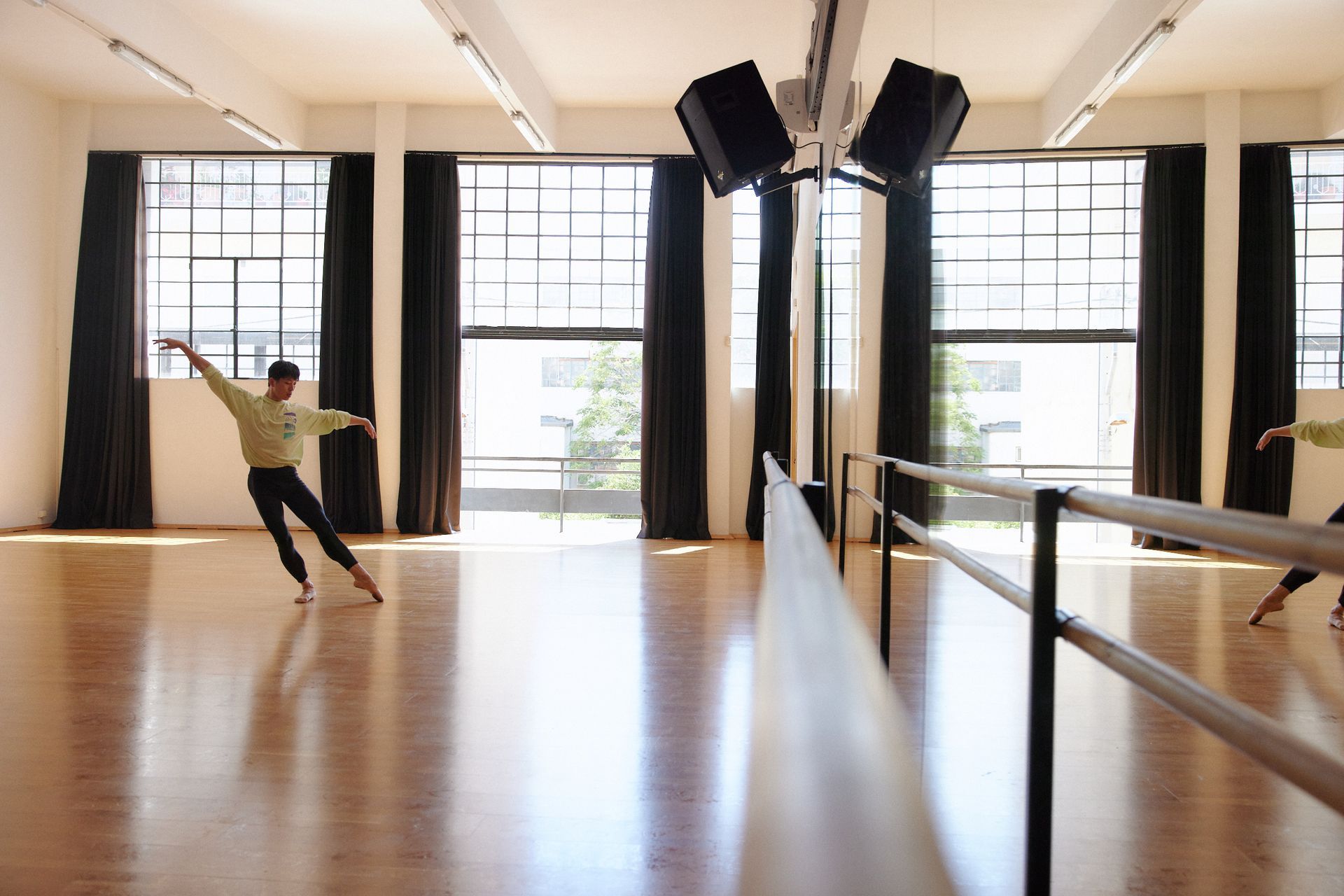 Dancer in a studio, with arm extended, reflecting in a long mirror. Light streams through windows, and a barre is visible.