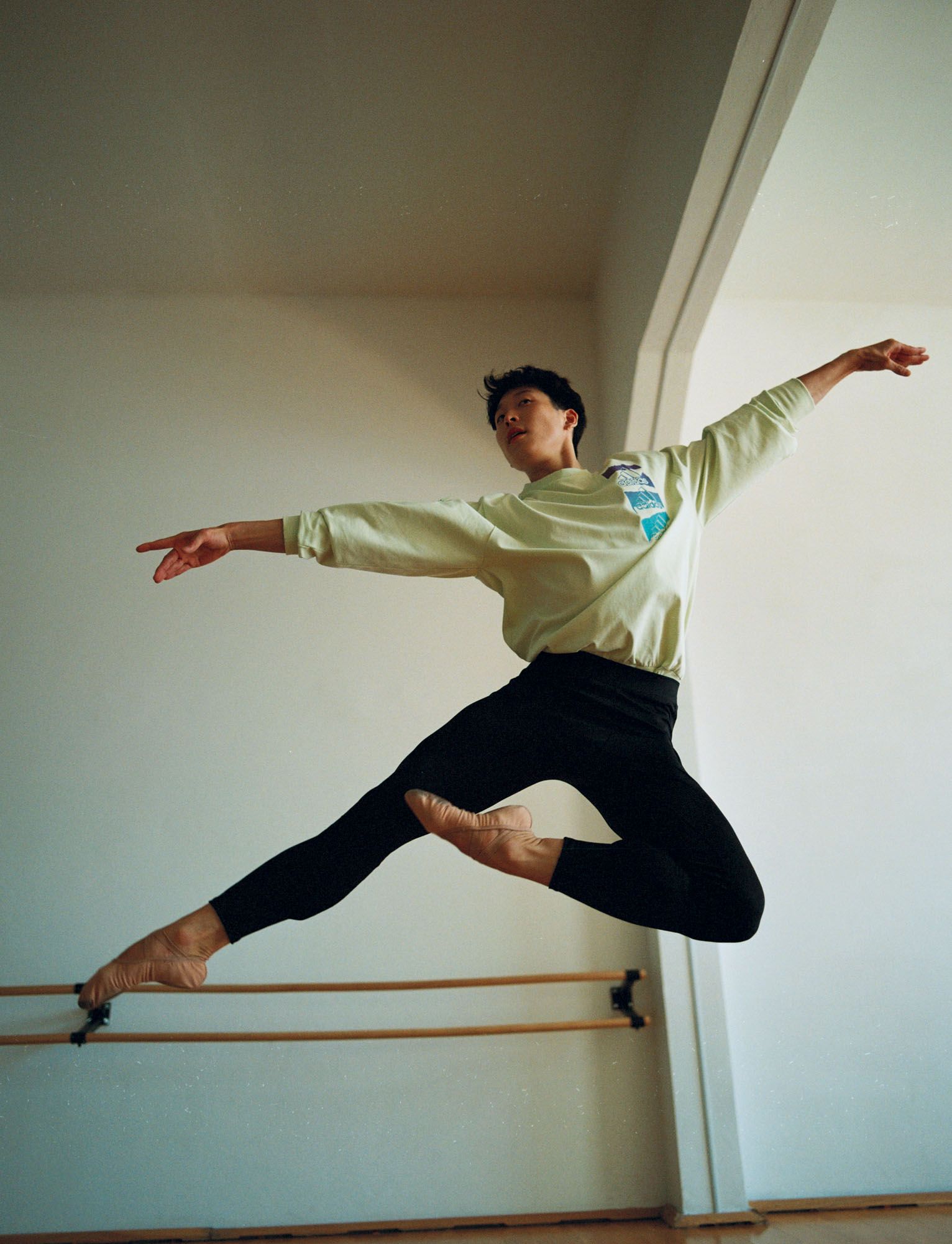 Dancer in mid-air leap, arms outstretched, black leggings, light-colored long-sleeve shirt, in front of a barre.