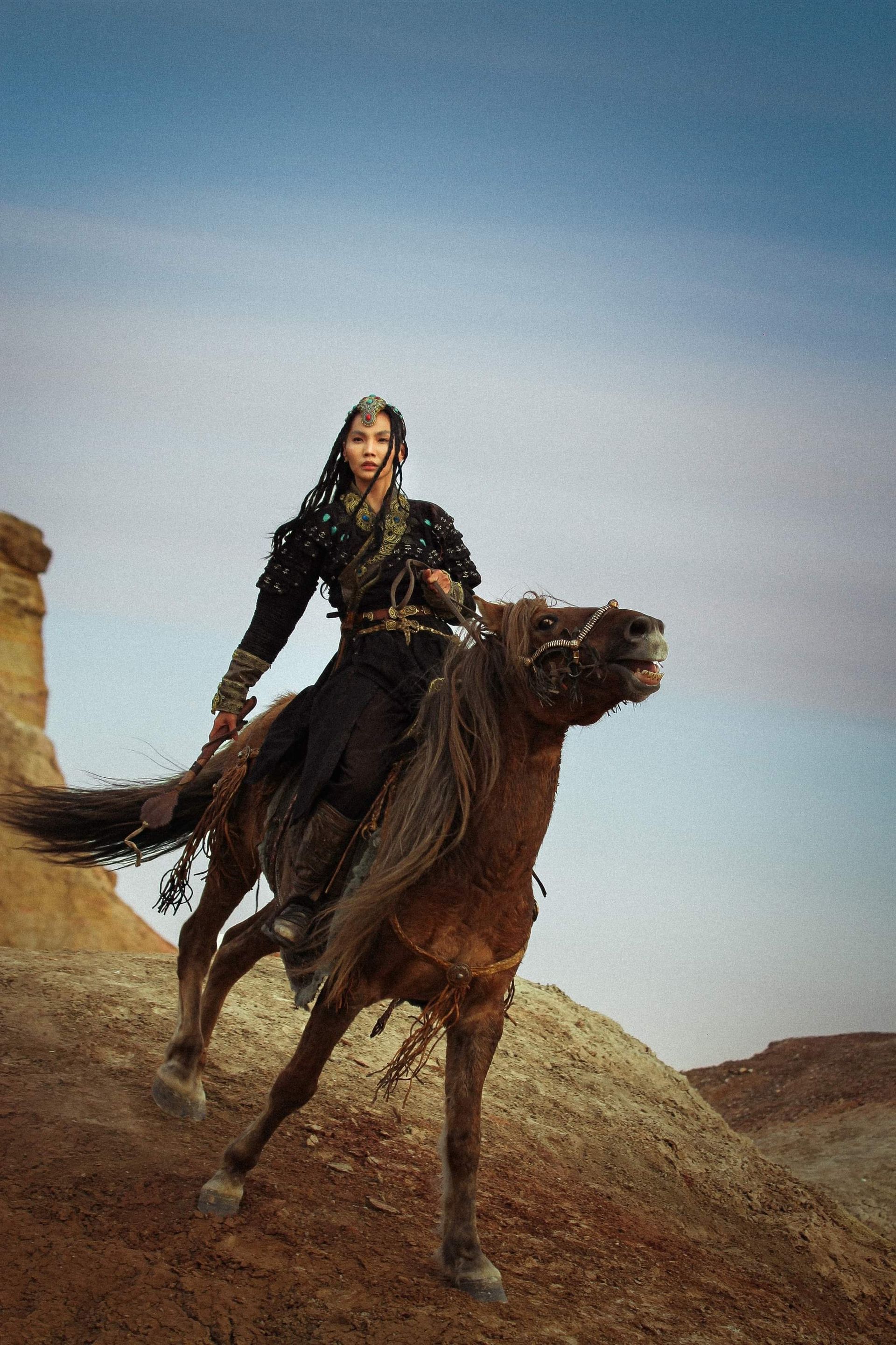 Woman riding a brown horse in a desert landscape. She's wearing dark clothing, and the horse is rearing its head.
