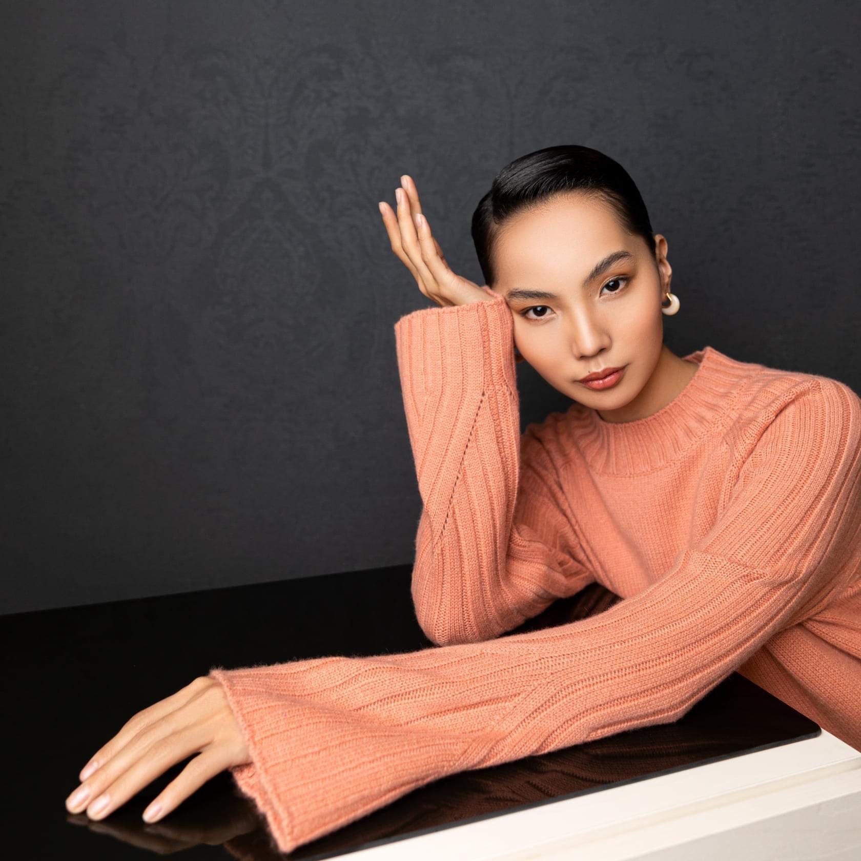 Woman leaning on a dark surface, wearing a peach sweater. She has dark hair and is looking towards the viewer.