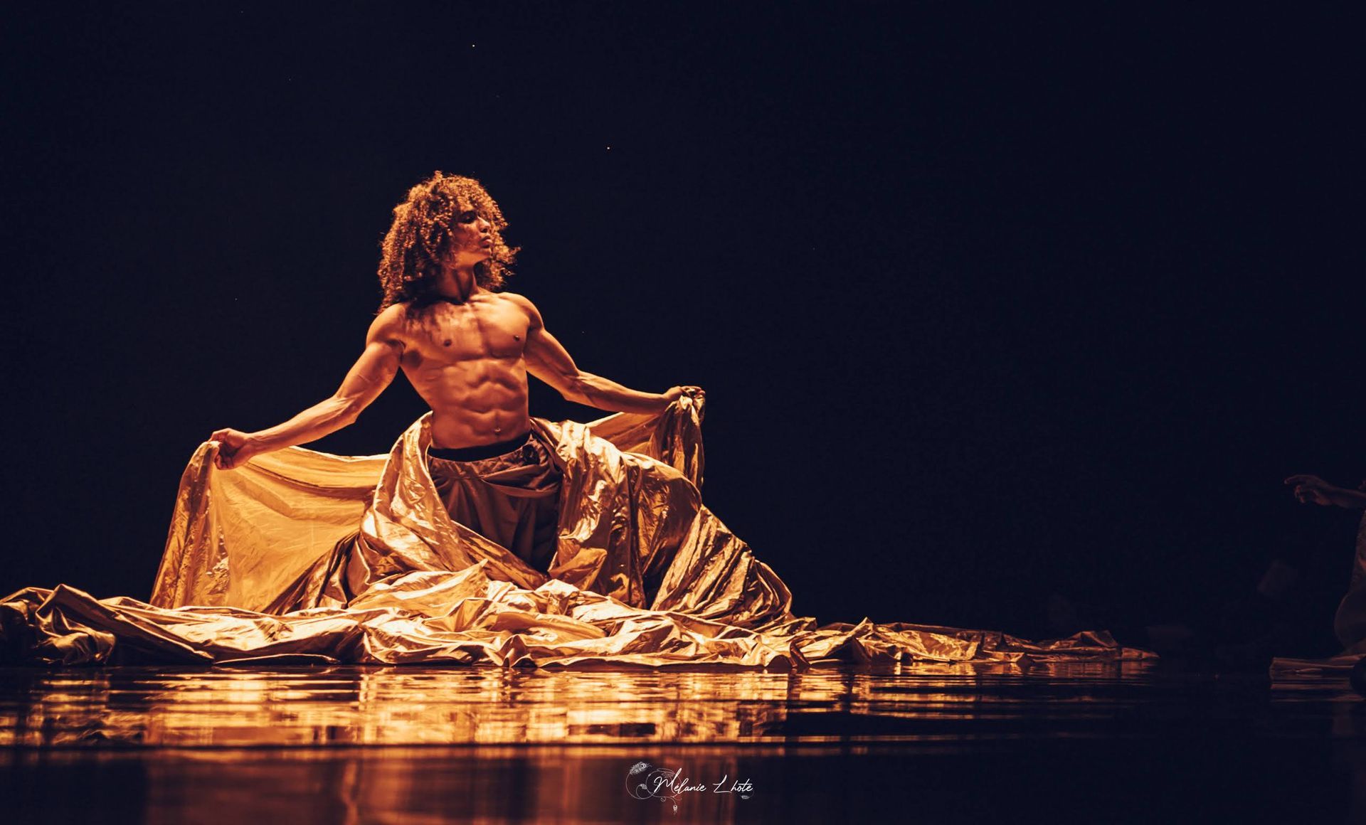 Male dancer in gold draped fabric, performing on reflective surface in dark theater.